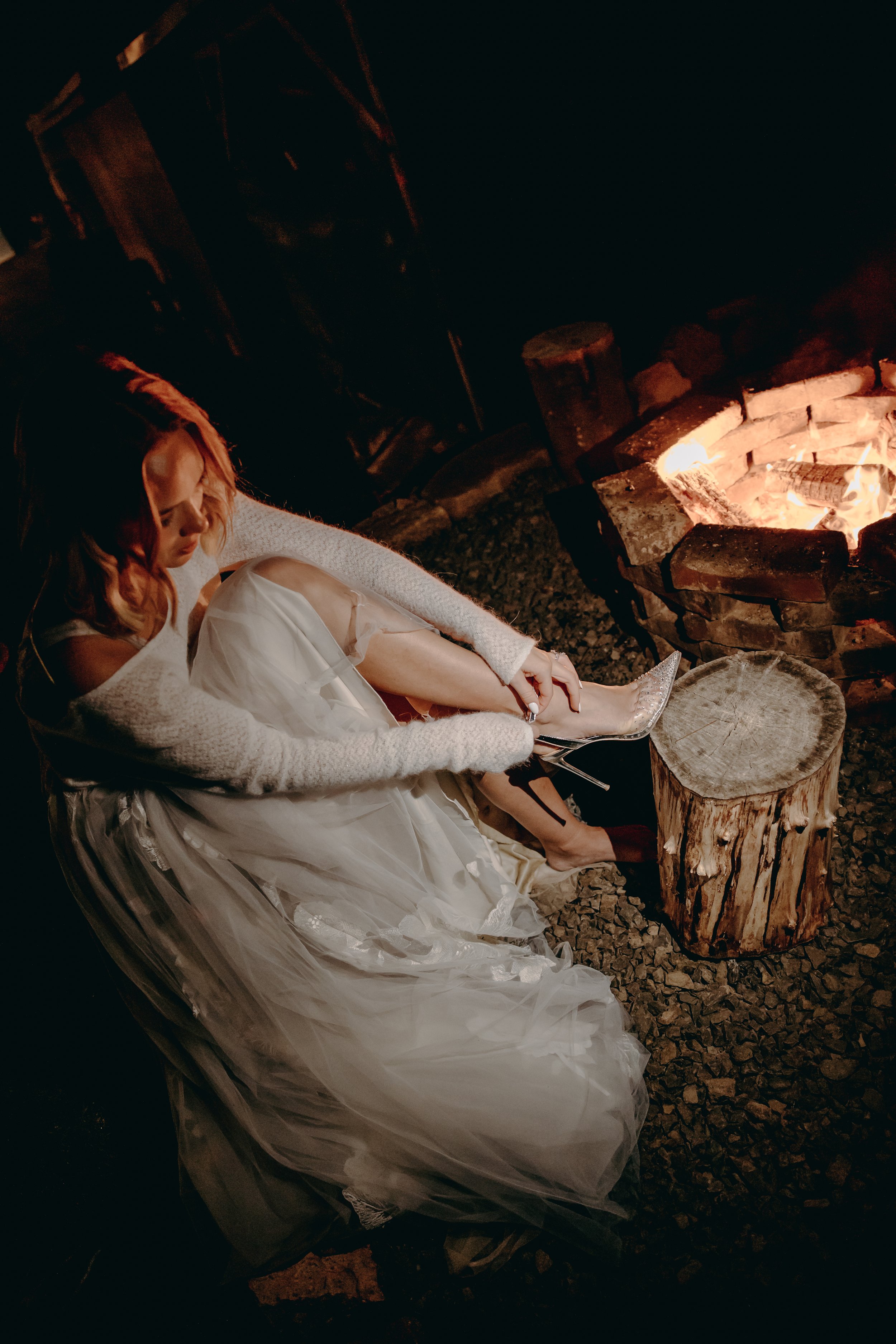 A woman in a wedding dress and a white cardigan sitting by a campfire in a dark outdoor setting.