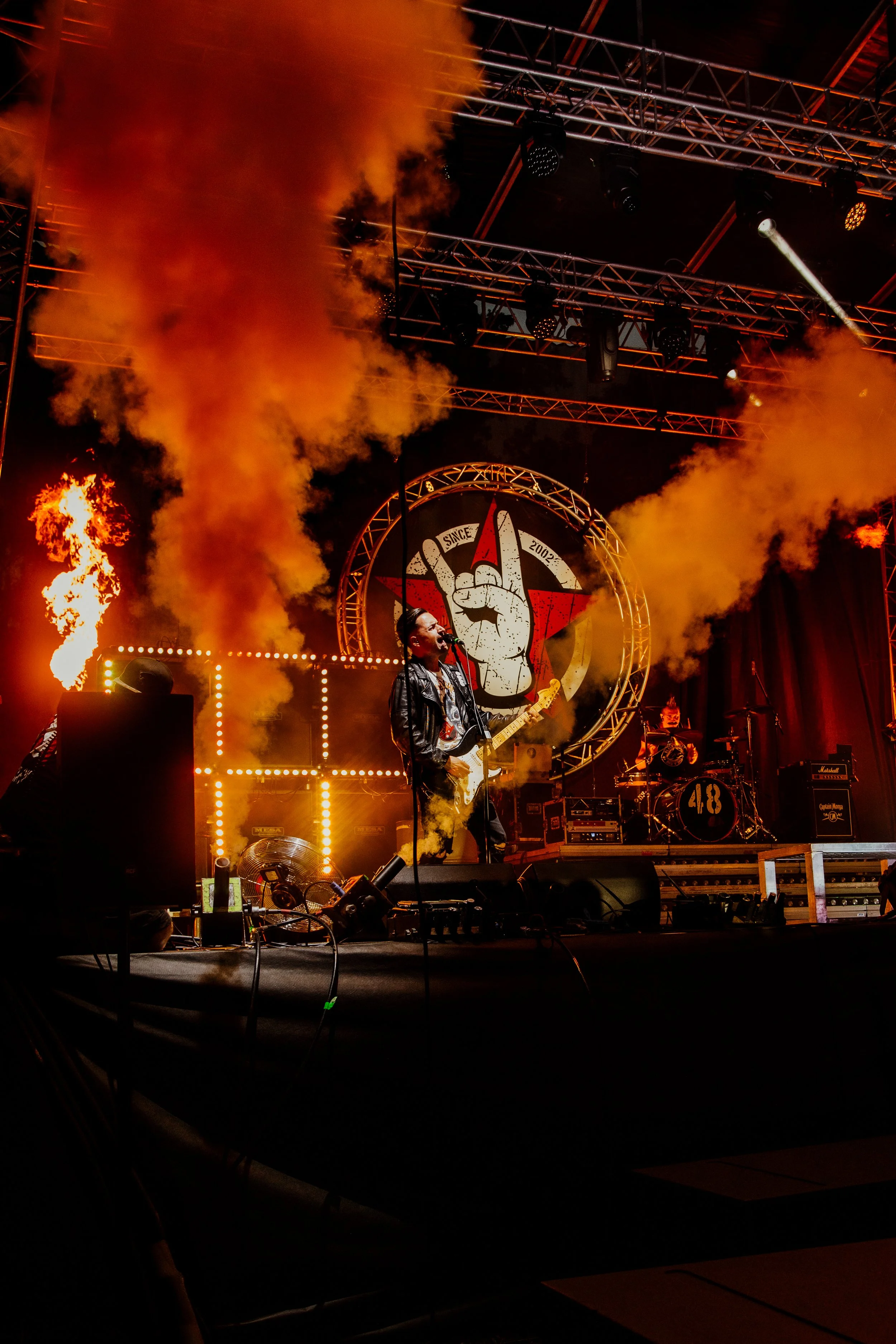 A live rock concert on stage with smoke and flames, a guitarist singing into a microphone, a drummer in the background, and a large logo featuring a hand making a rock and roll gesture behind them.