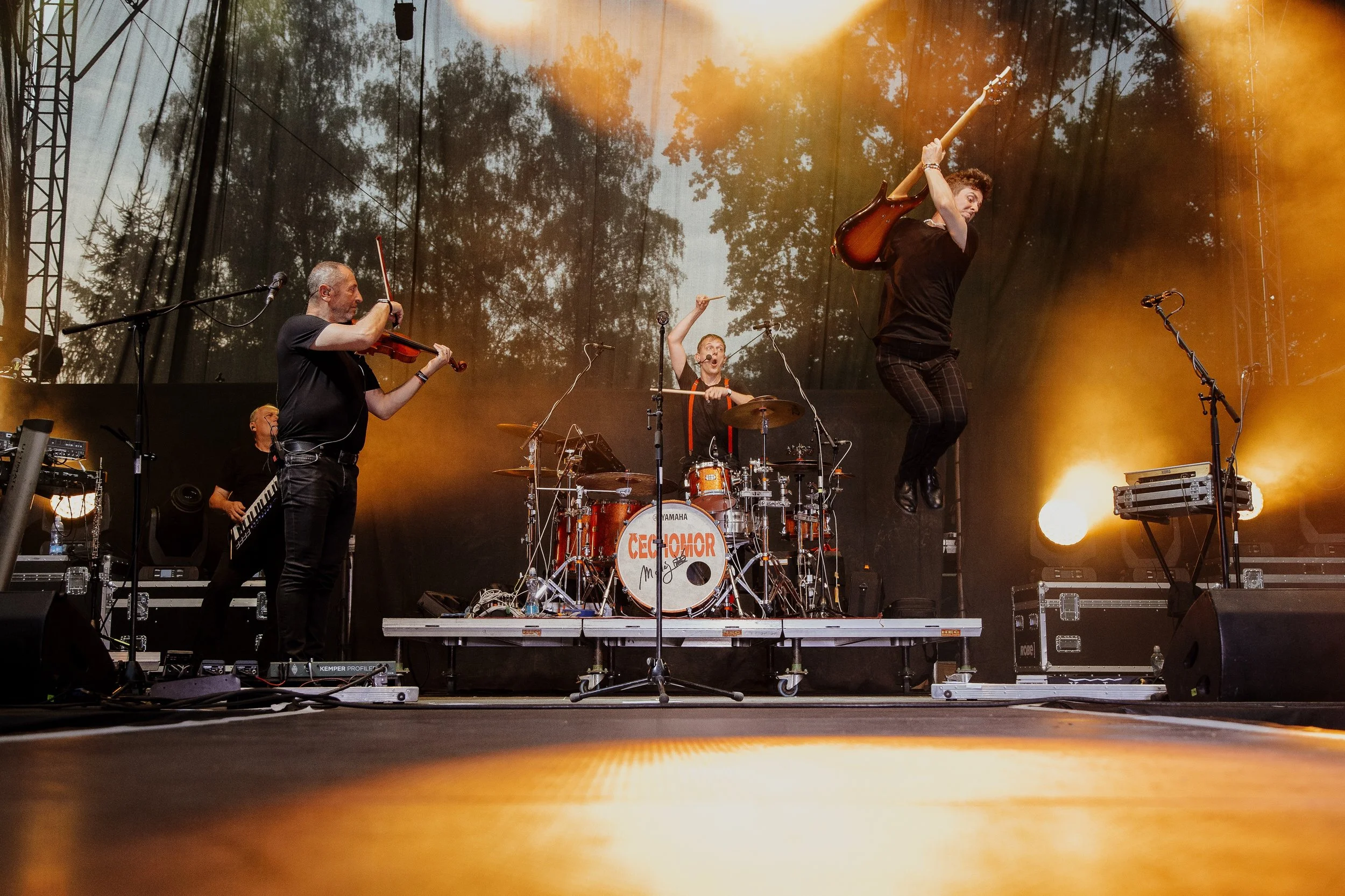 Musicians performing on stage with one playing the violin, one on drums, and one jumping with an electric guitar. Stage is lit with yellow lighting, and trees can be seen in the background.