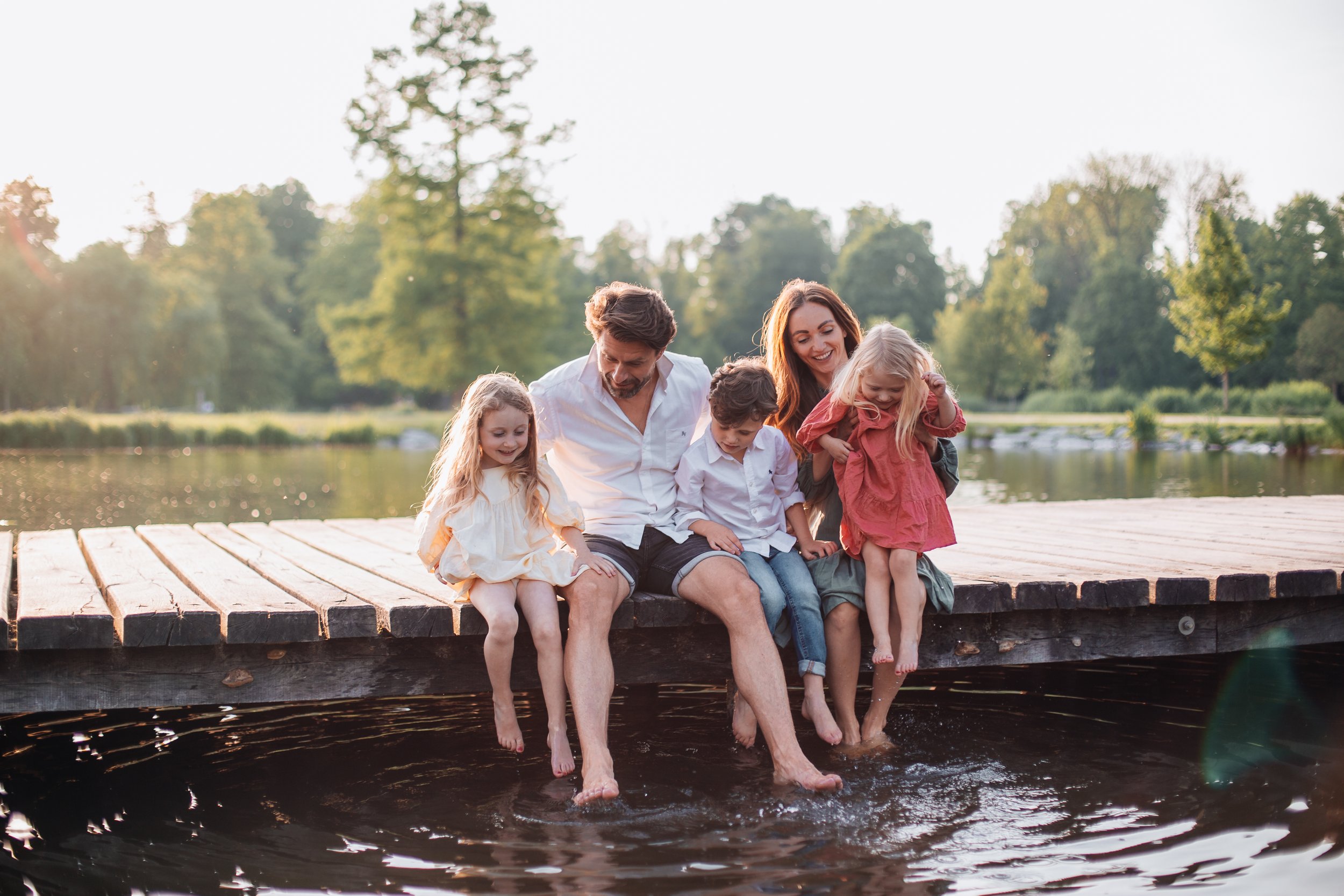 Family of six sitting on a wooden dock with their feet in the water, enjoying a sunny day by the lake, surrounded by trees.