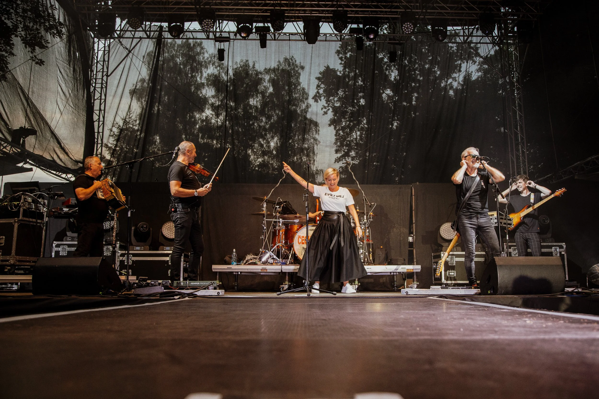 Band performing on stage outdoors, with five members, including a woman with her arms raised, while others play instruments such as violin, accordion, bass, and guitar.