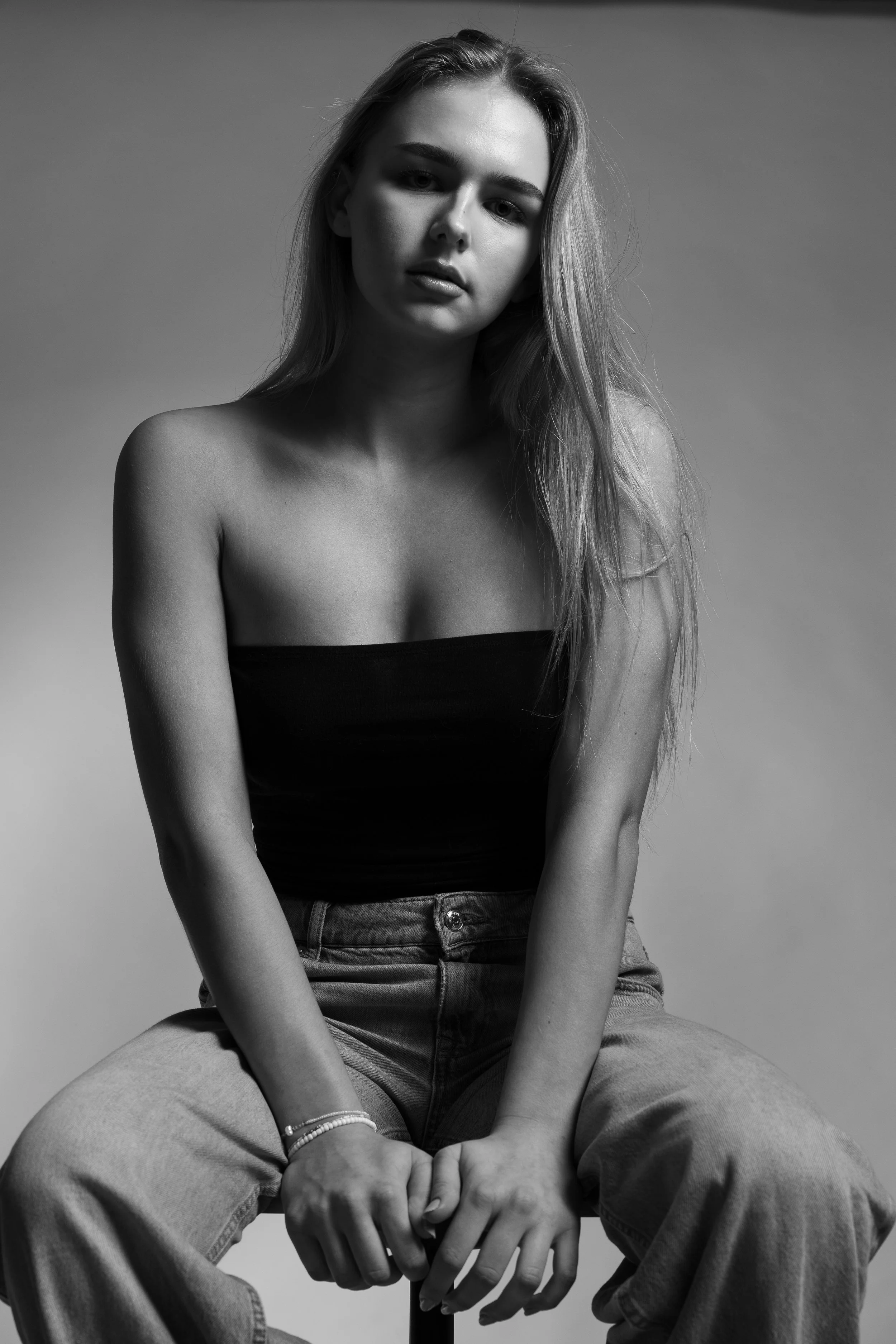 Black and white portrait of a young woman with long hair wearing a strapless top and jeans, sitting in a relaxed pose against a plain background.