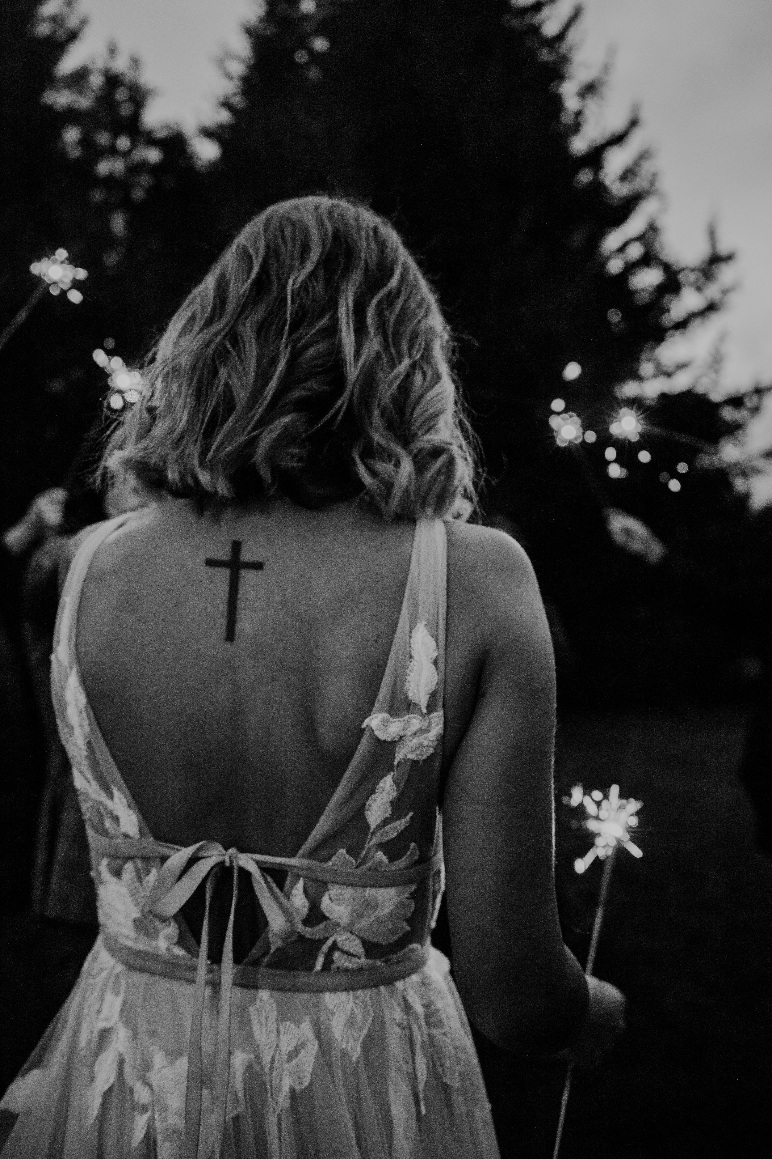 A woman with shoulder-length wavy hair wearing a sleeveless dress with floral embroidery, holding a sparkler, with a large cross tattoo on her upper back, outdoors during twilight.