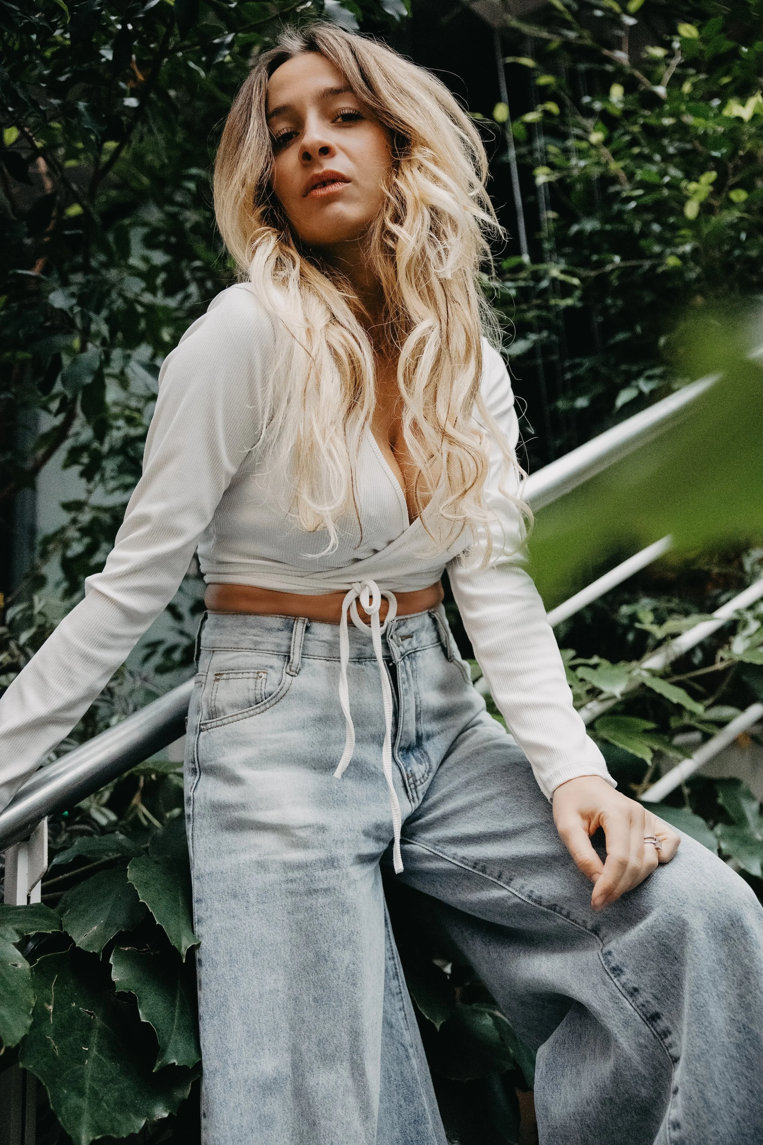 A young woman with long, blonde, curly hair standing outdoors among green foliage. She is wearing a white cropped top with long sleeves and high-waisted, loose-fitting light-wash jeans. She is leaning against a metal railing, looking at the camera with a relaxed expression.