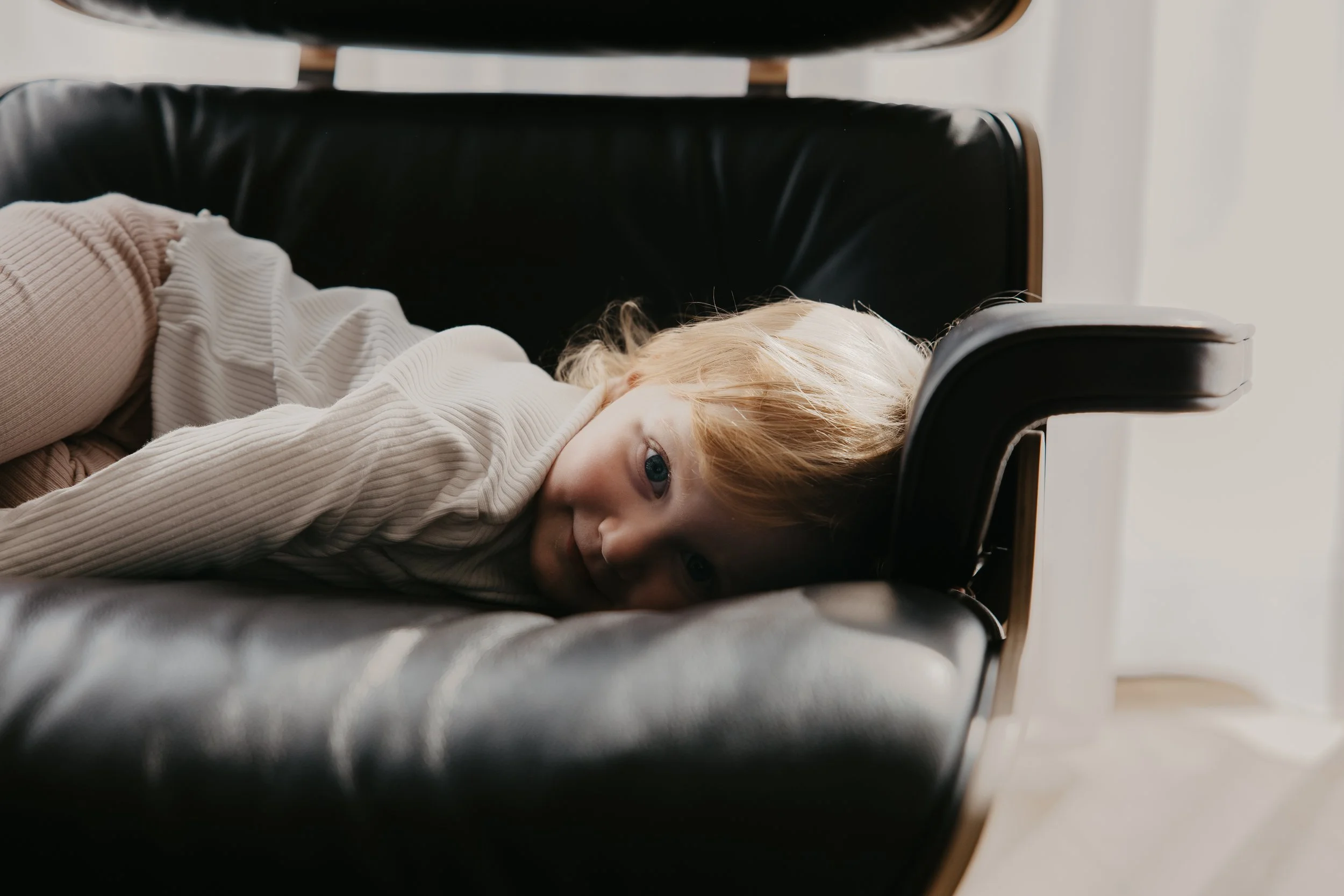 A young girl with blonde hair and blue eyes lying down with her head resting on a black leather chair and looking at the camera.