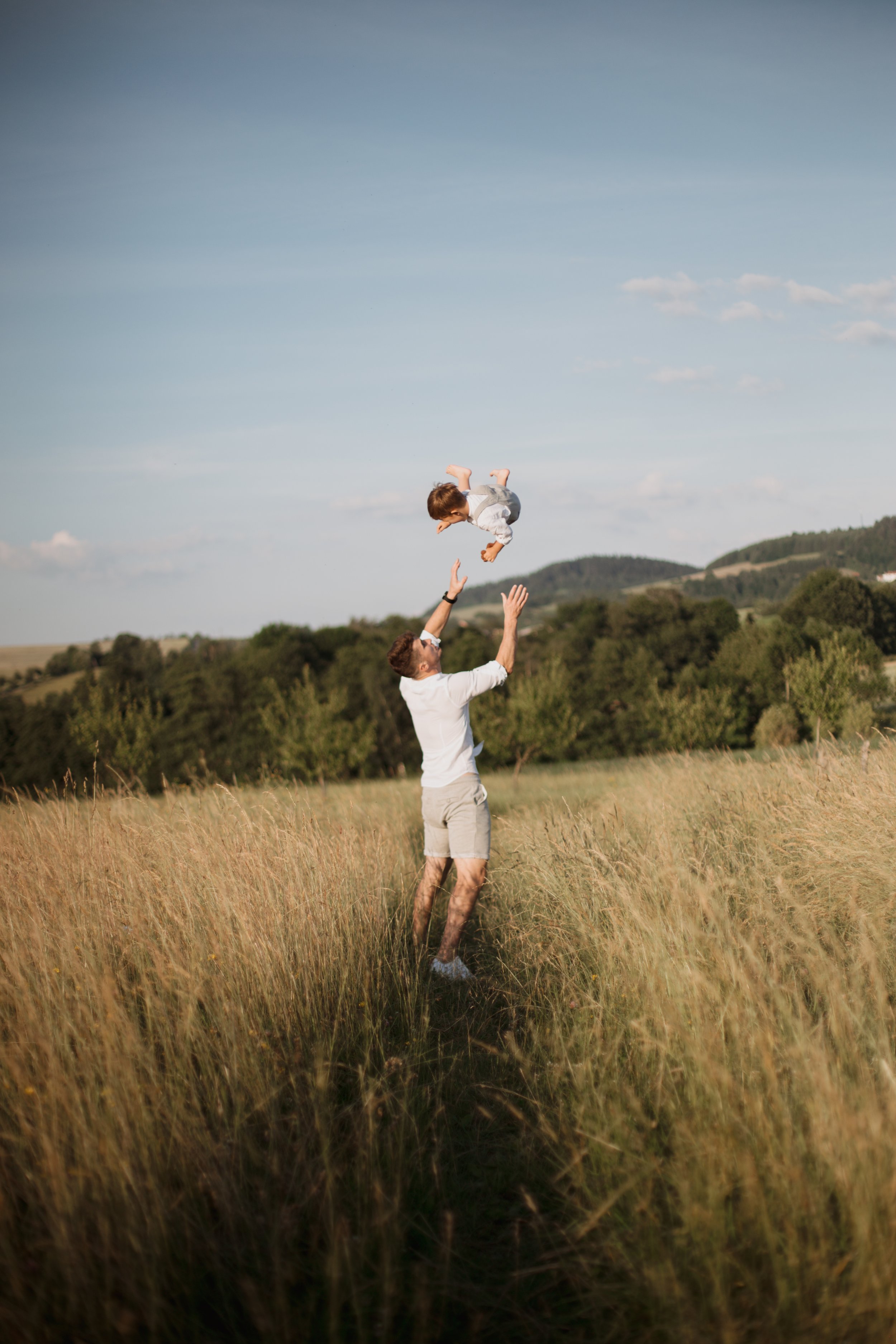 A man is throwing a young boy into the air in a grassy field under a blue sky.