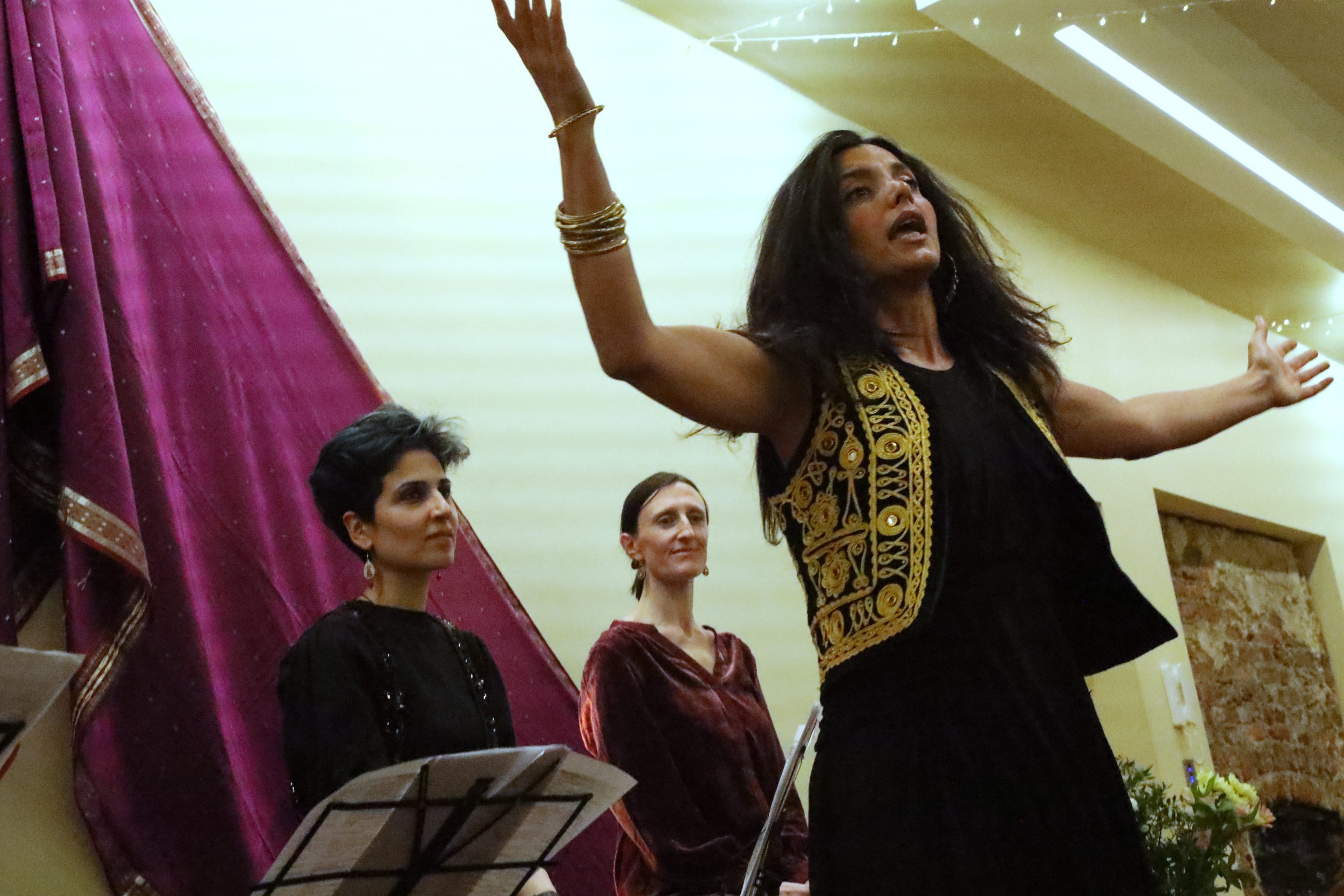 A woman in a black and gold embroidered vest raises her arms expressively while two others, seated, observe her with musical notes in front of them. They are indoors with a maroon fabric backdrop.