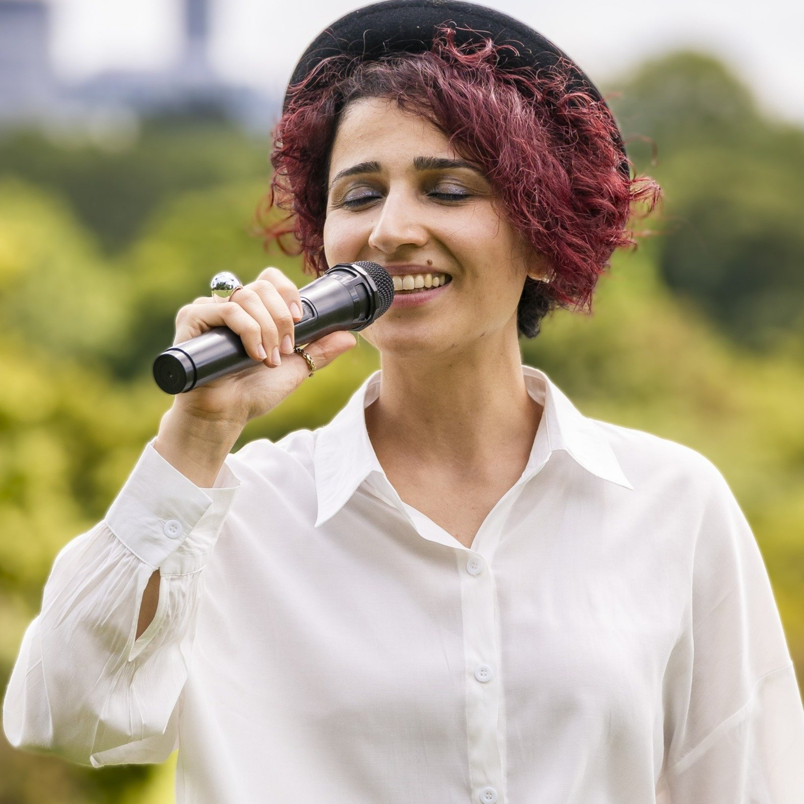 Woman singing outdoors with a microphone, wearing a white shirt and black hat, surrounded by greenery.