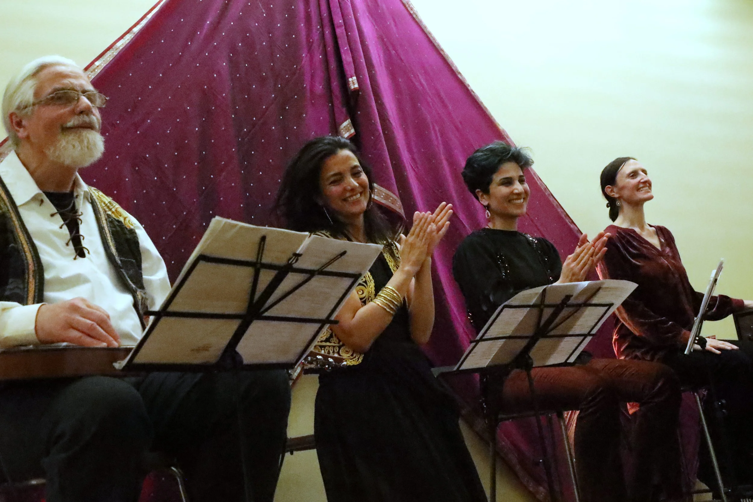 Four musicians sitting and smiling, with sheet music stands in front. A large maroon drapery is in the background.