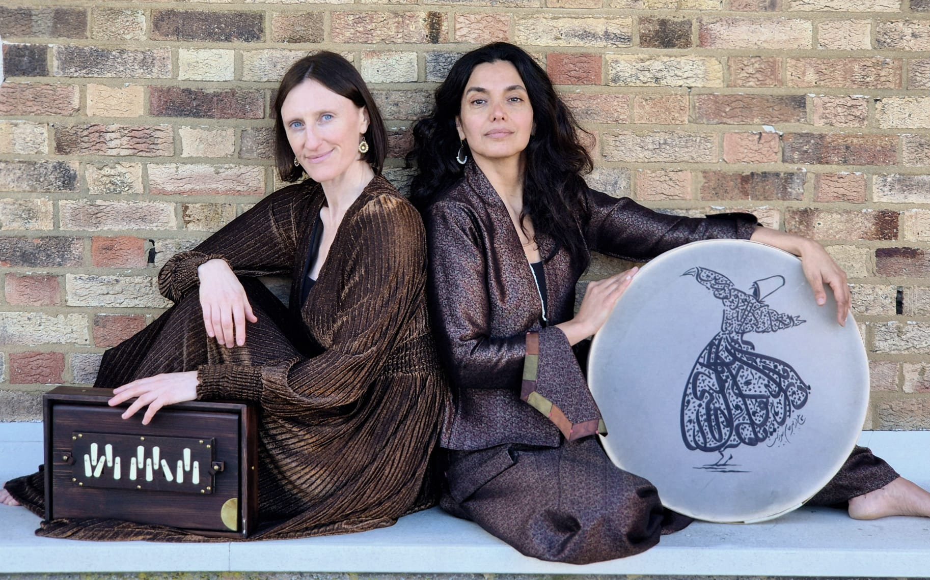 Two women sitting against a brick wall, one holding a wooden harmonium, the other holding a drum with intricate design.