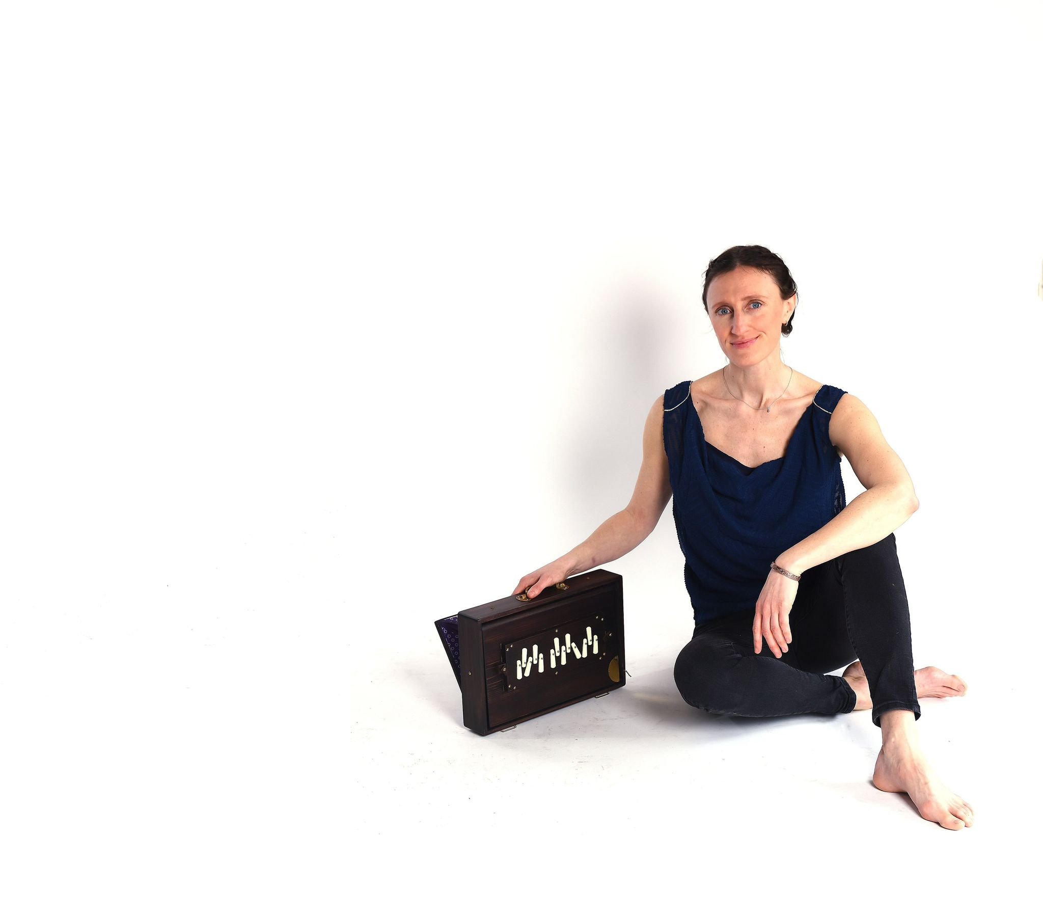 A woman sitting on the floor with a harmonium on her lap against a white background. She is barefoot and dressed in a blue top and black pants.