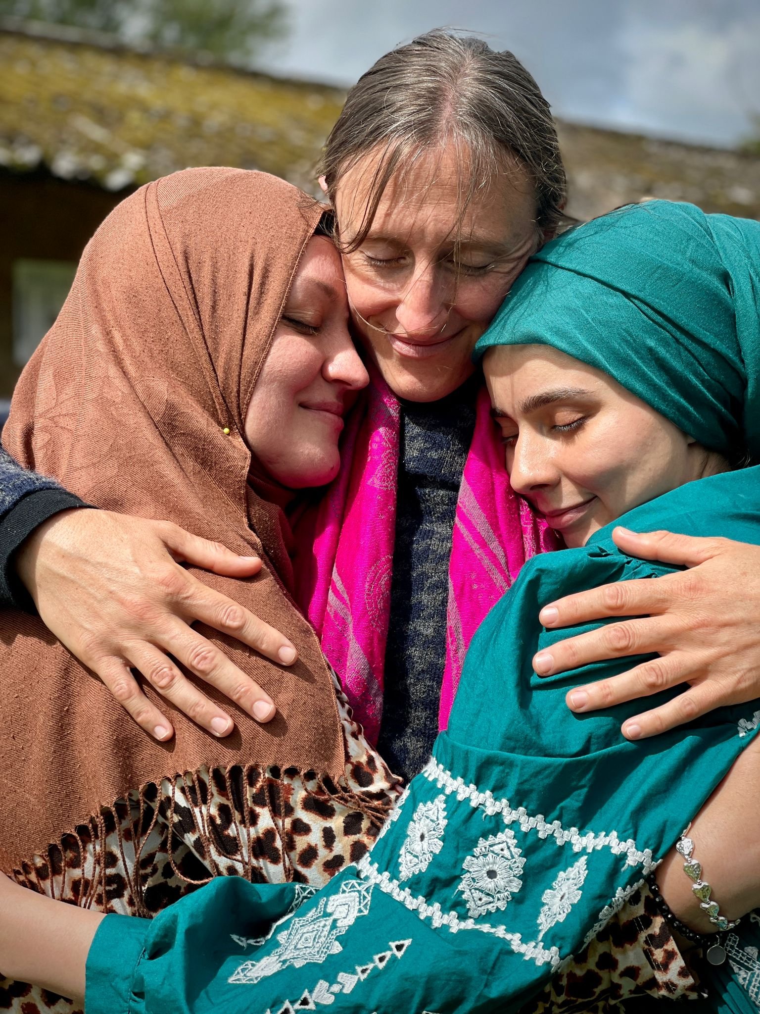 Three people hugging closely with smiles, wearing colorful headscarves and shawls; background of a roof and sky.