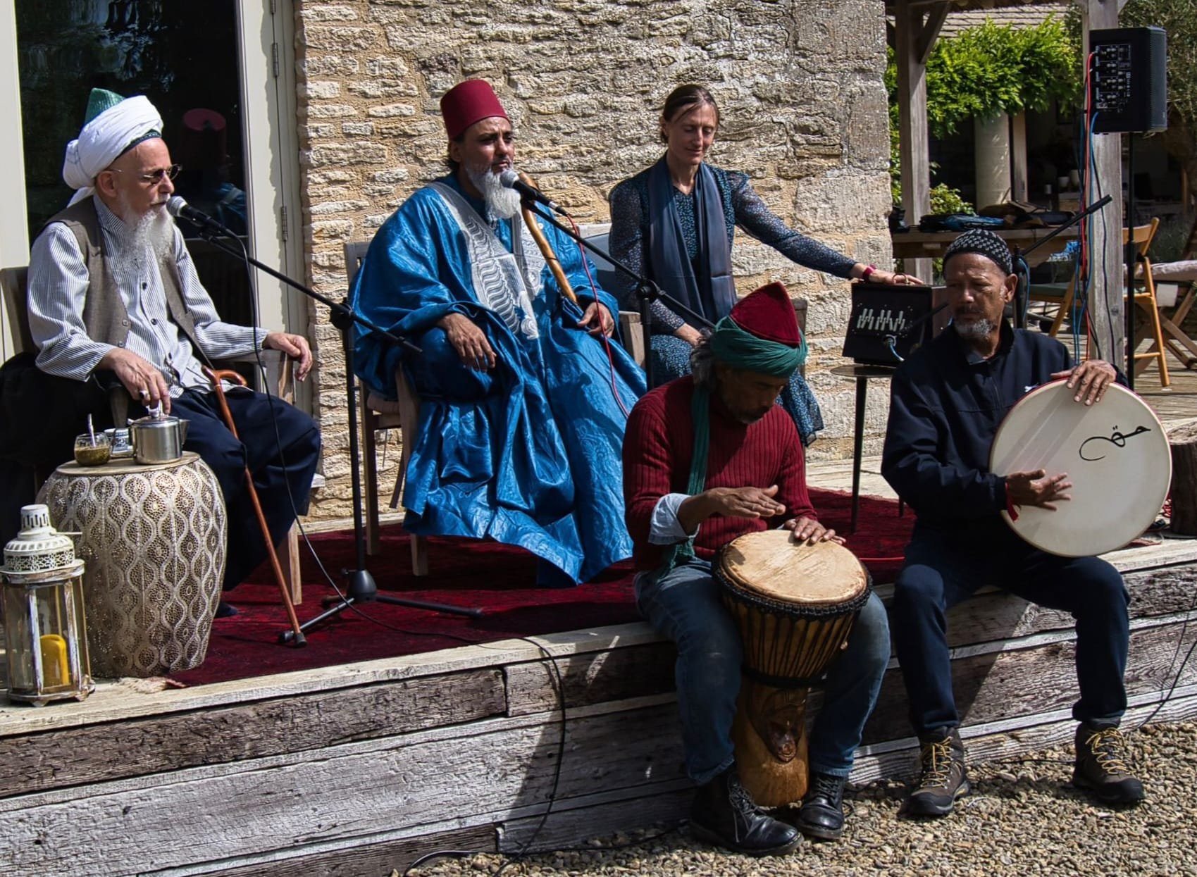 Group of musicians performing outdoors, with traditional instruments and diverse attire, set against a stone wall backdrop.