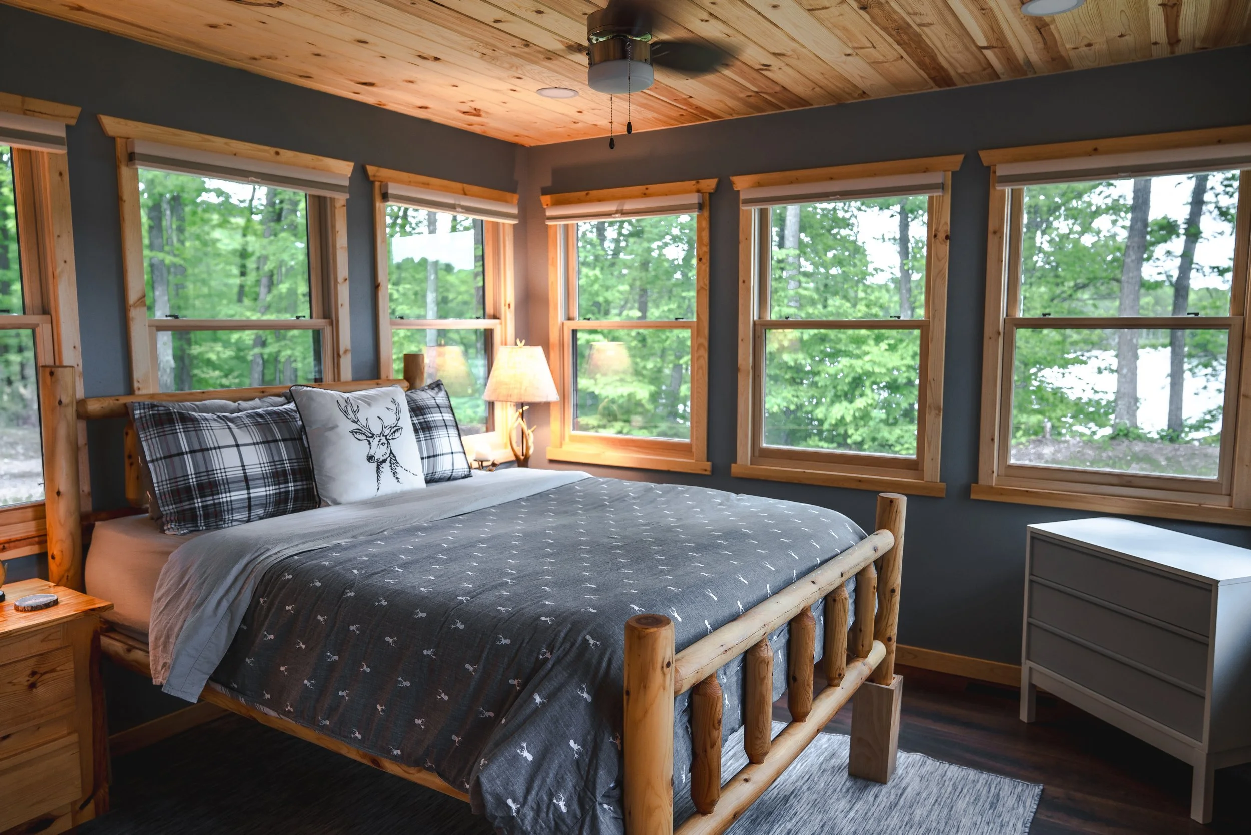 Cozy bedroom with wooden bed frame, plaid pillows, a decorative pillow with a deer, surrounded by large windows showing green trees outside, wooden ceiling, and a nightstand with a lamp.