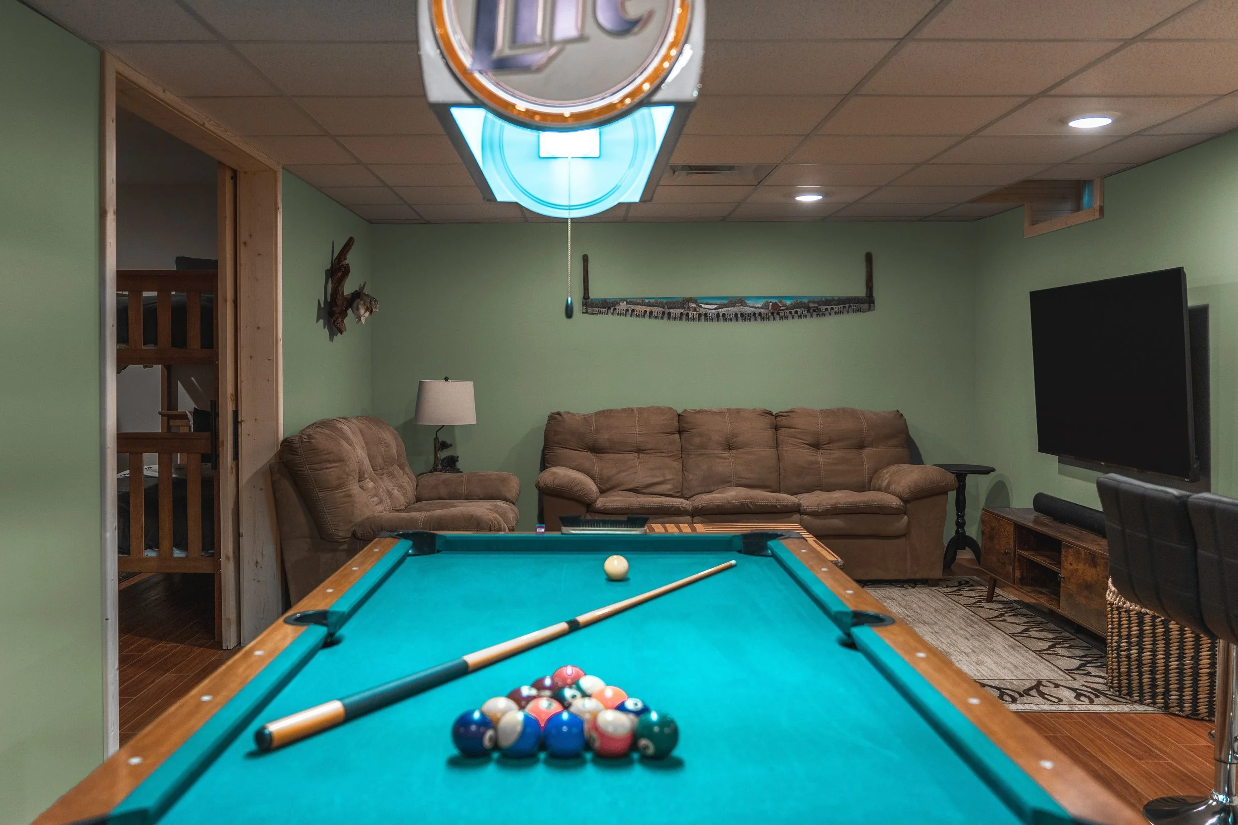 Pool table with billiard balls and cue stick in a basement game room, featuring a brown sofa, a flat-screen TV, and green walls.