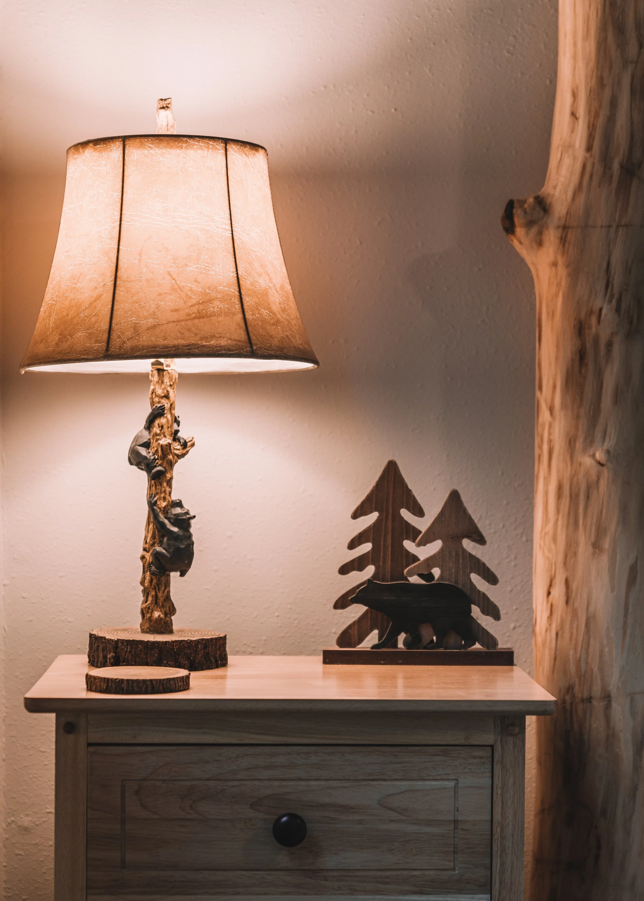 A wooden table with a rustic lamp featuring a branch and bear design, two circular wood coasters, and a decorative wooden bear and pine trees sculpture, with part of a wood beam visible.
