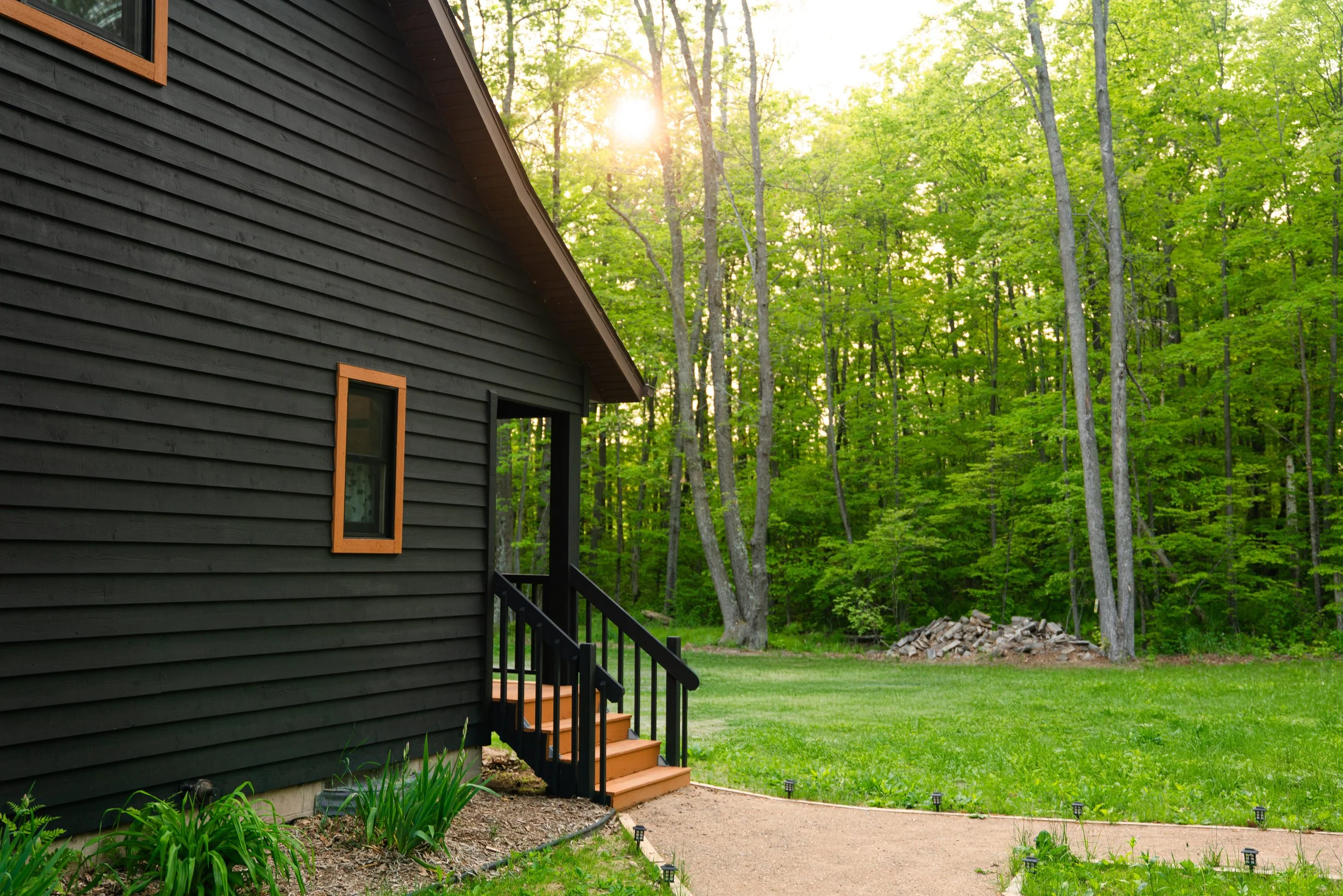 Side of a black house with orange-framed window, small porch with steps, greenery, and a dense forest with tall trees in the background, sunny sky.