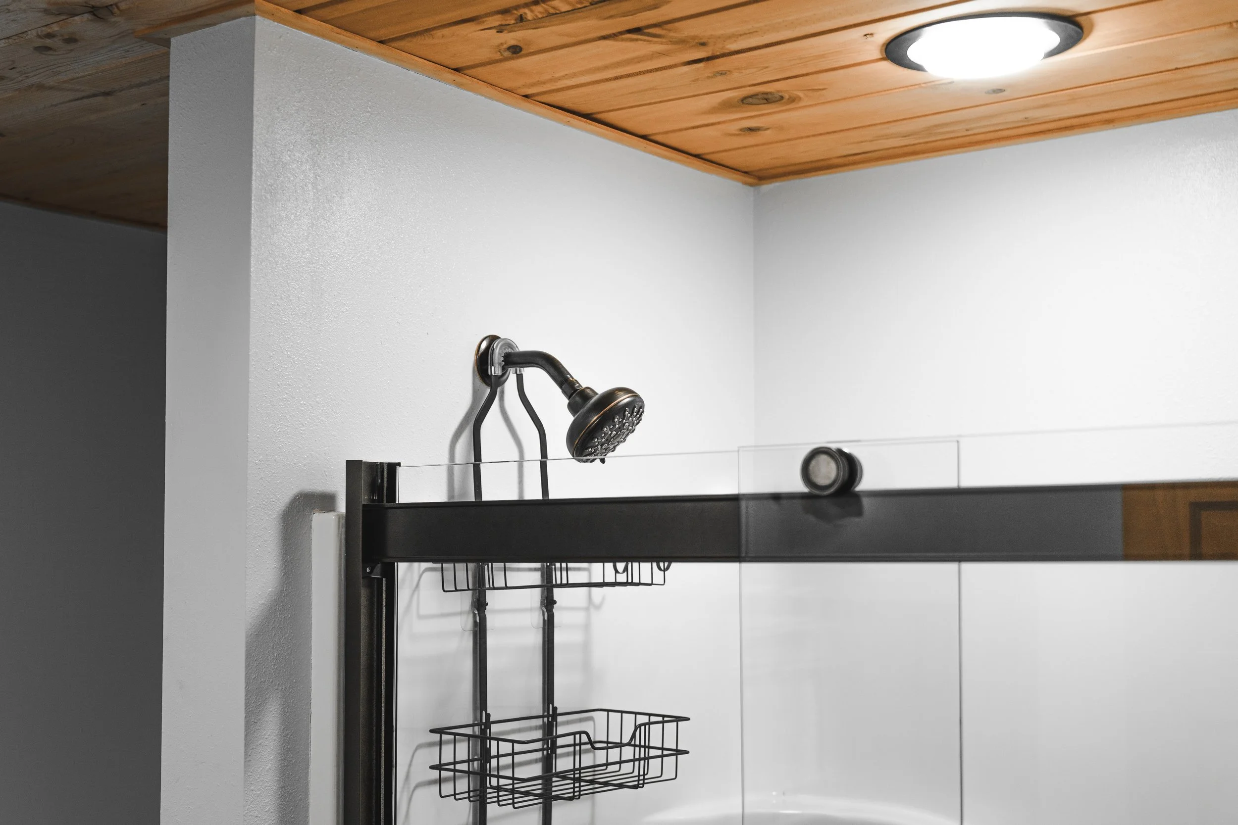 Shower area with a black showerhead, wall-mounted shelf with a glass panel, and a wooden ceiling with a round ceiling light.
