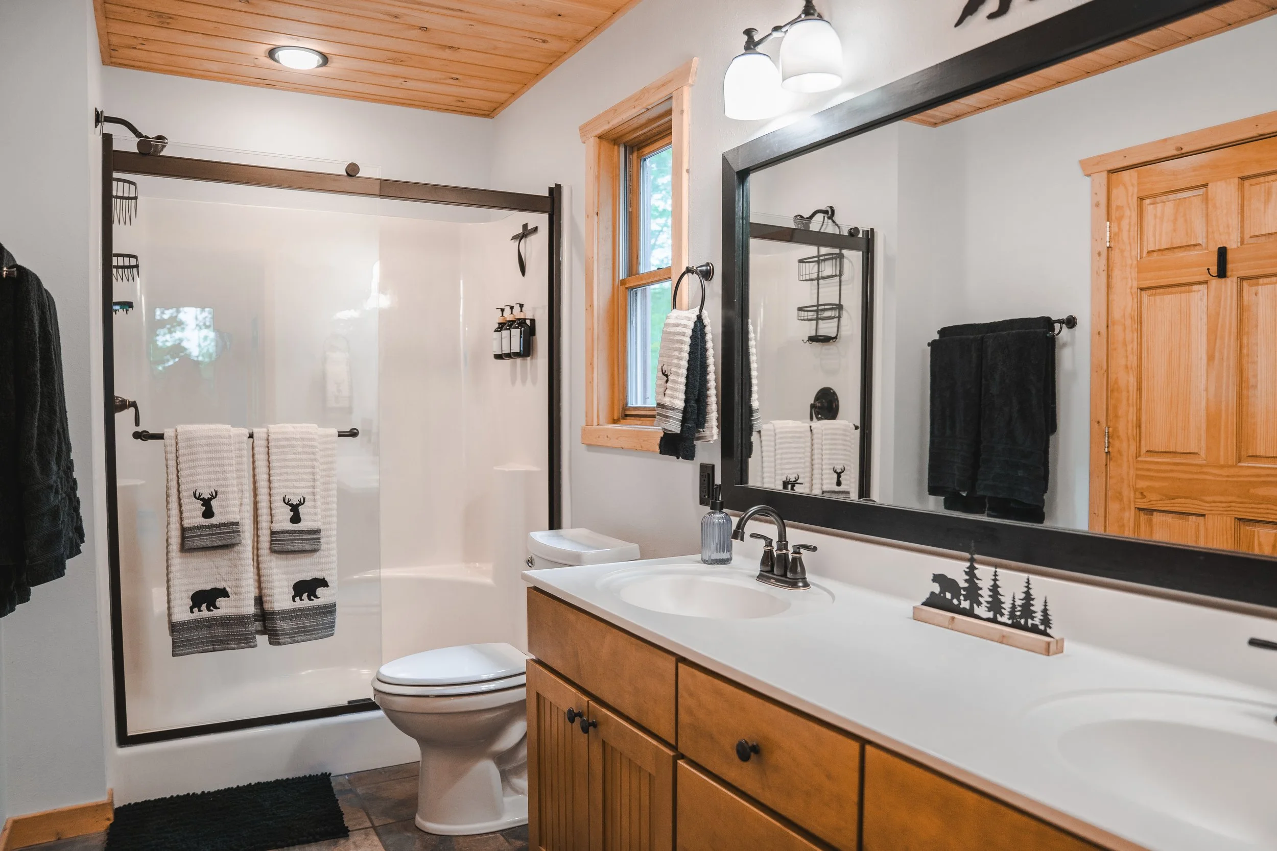 Bathroom with a wooden ceiling, white walls, a double sink vanity with a large mirror, a window, a glass-enclosed shower with white and black towels, and wood accents.