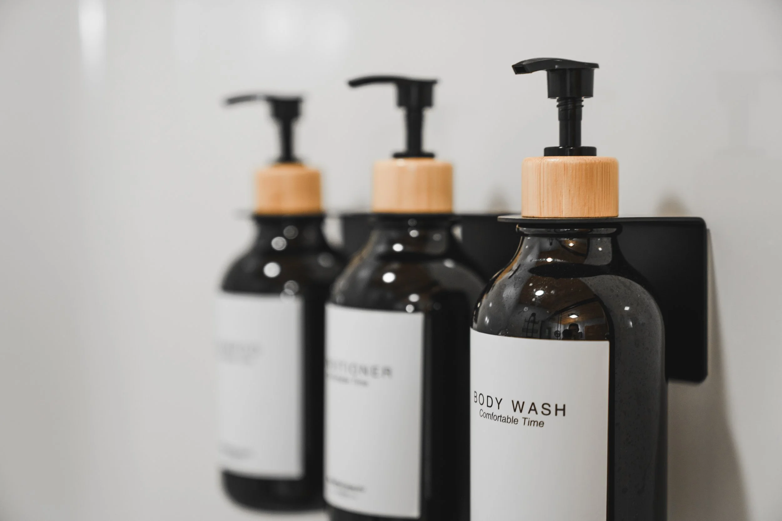 Three black bottles of body wash with wooden pump tops mounted on a black wall-mounted dispenser, with a blurred background.