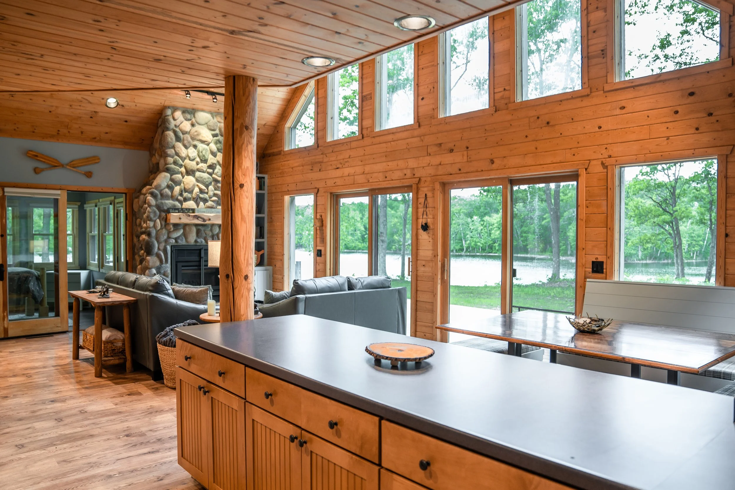 Interior view of a wooden cabin with large windows overlooking a lake and lush greenery, featuring a stone fireplace, leather sofas, a dining area, and decorative canoe paddles on the wall.