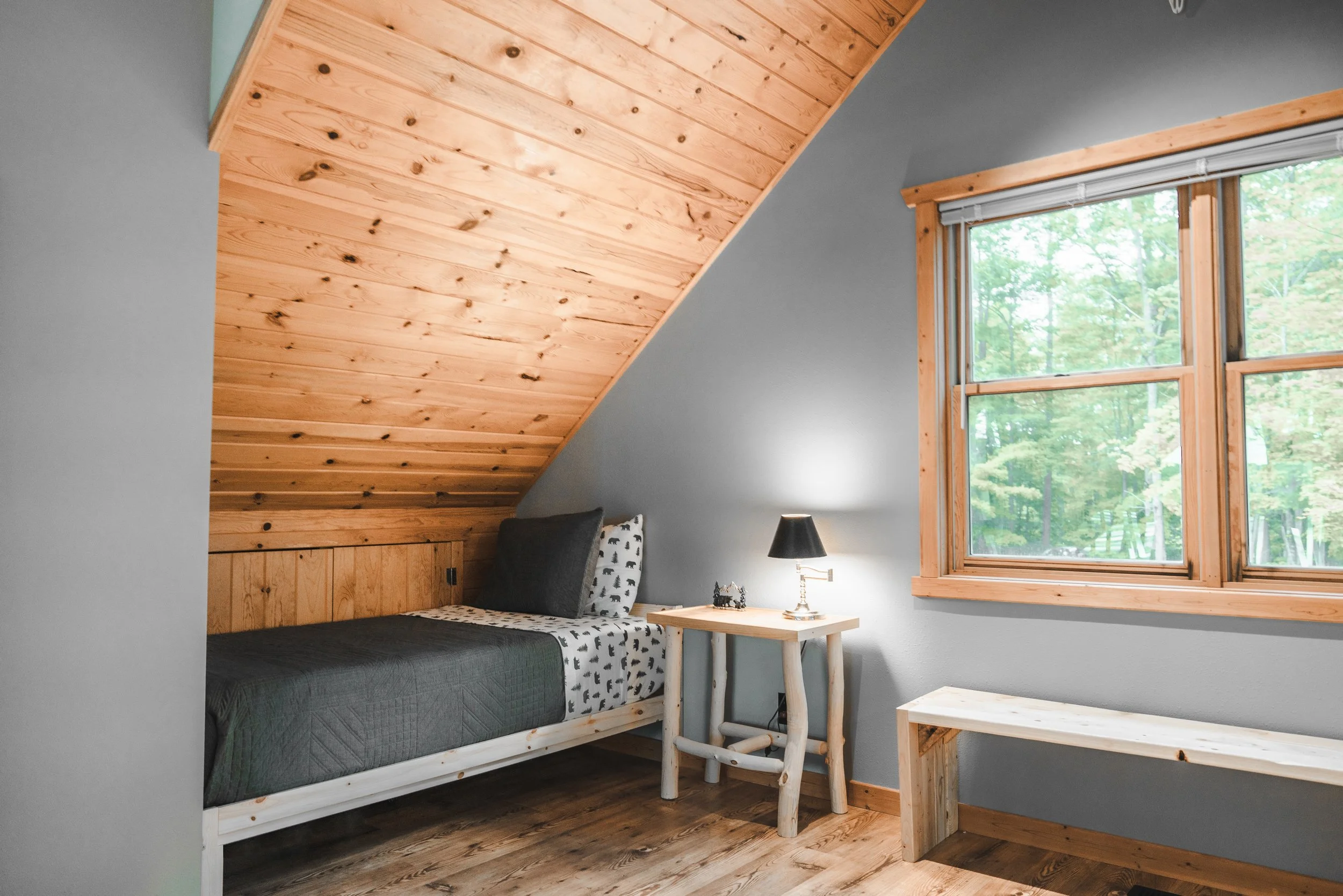 A cozy bedroom with a sloped wooden ceiling, a single bed with black and white bedding, a small wooden side table with a black lamp, and a large window showing green trees outside.