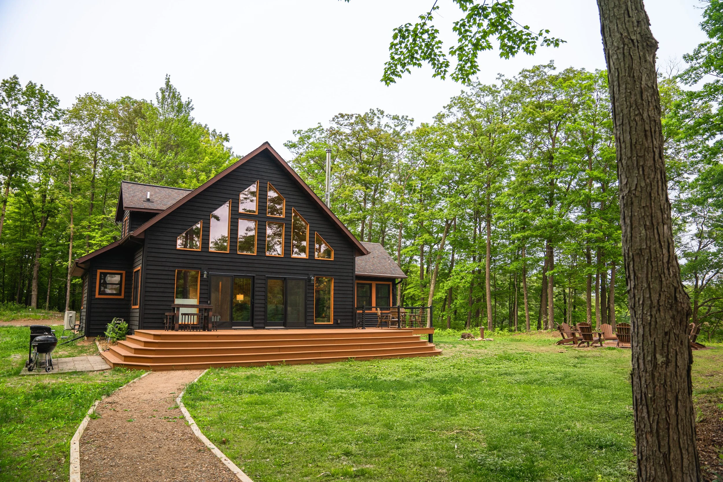 Black wooden house with large windows and a wooden deck, surrounded by green grass, trees, and outdoor seating in a forested area.