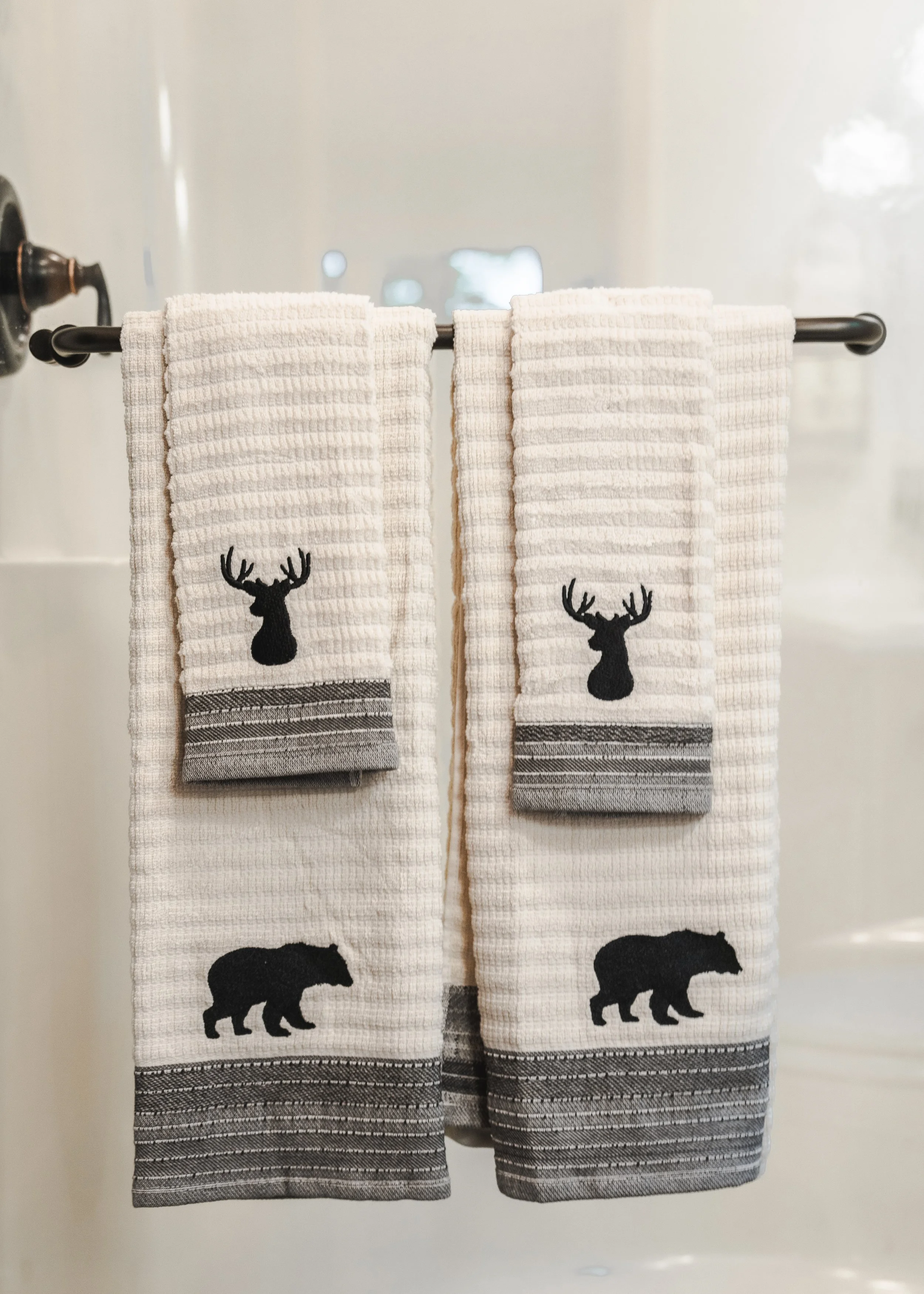 Two white towels with black silhouettes of a deer and a bear, hanging on a black towel bar in front of a white background.