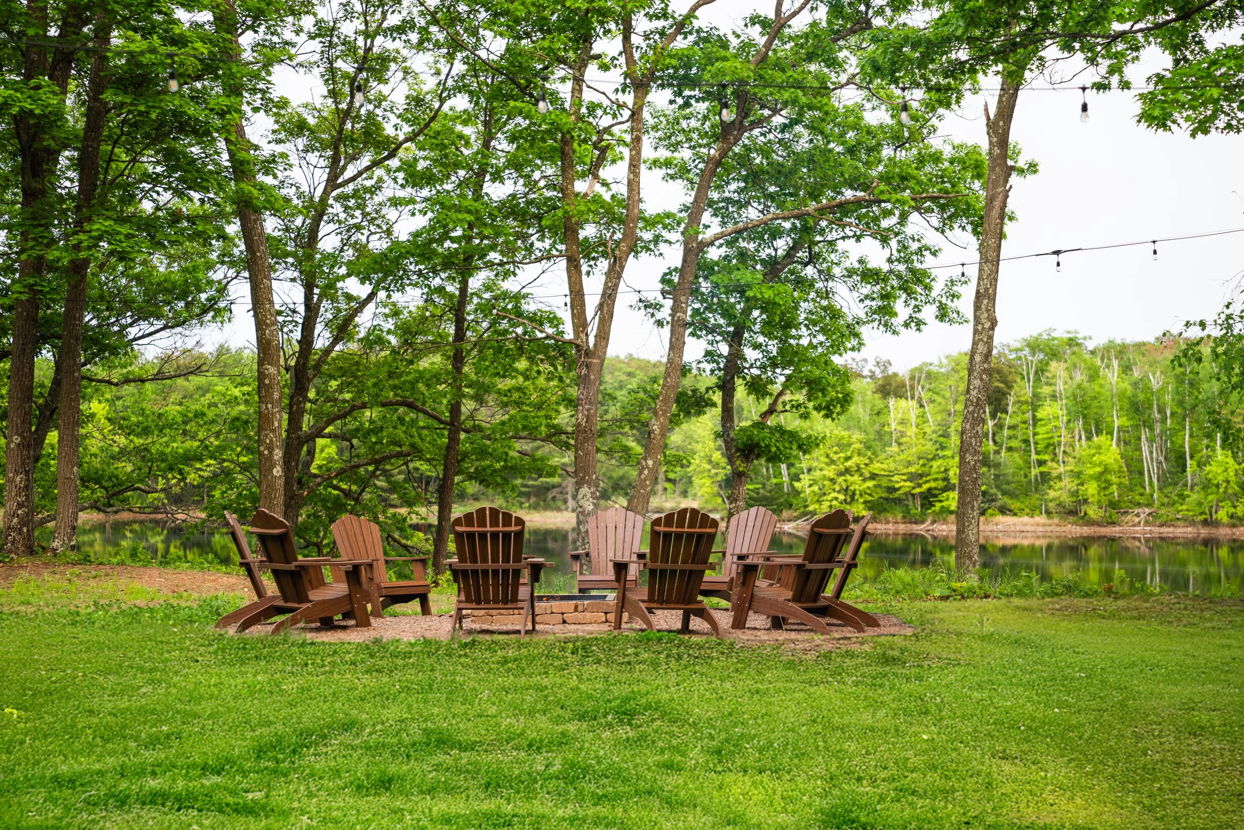 Six brown Adirondack chairs arranged in a circle around a fire pit on a grassy area by a river, with tall trees and string lights overhead.