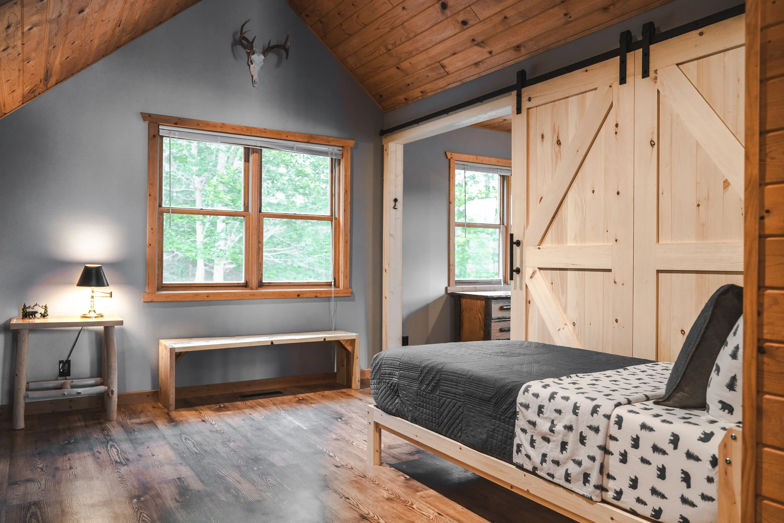 Rustic bedroom with gray walls, wooden floors, a bed with black and patterned bedding, a wooden sliding barn door, and natural light coming through two windows with views of green trees outside.