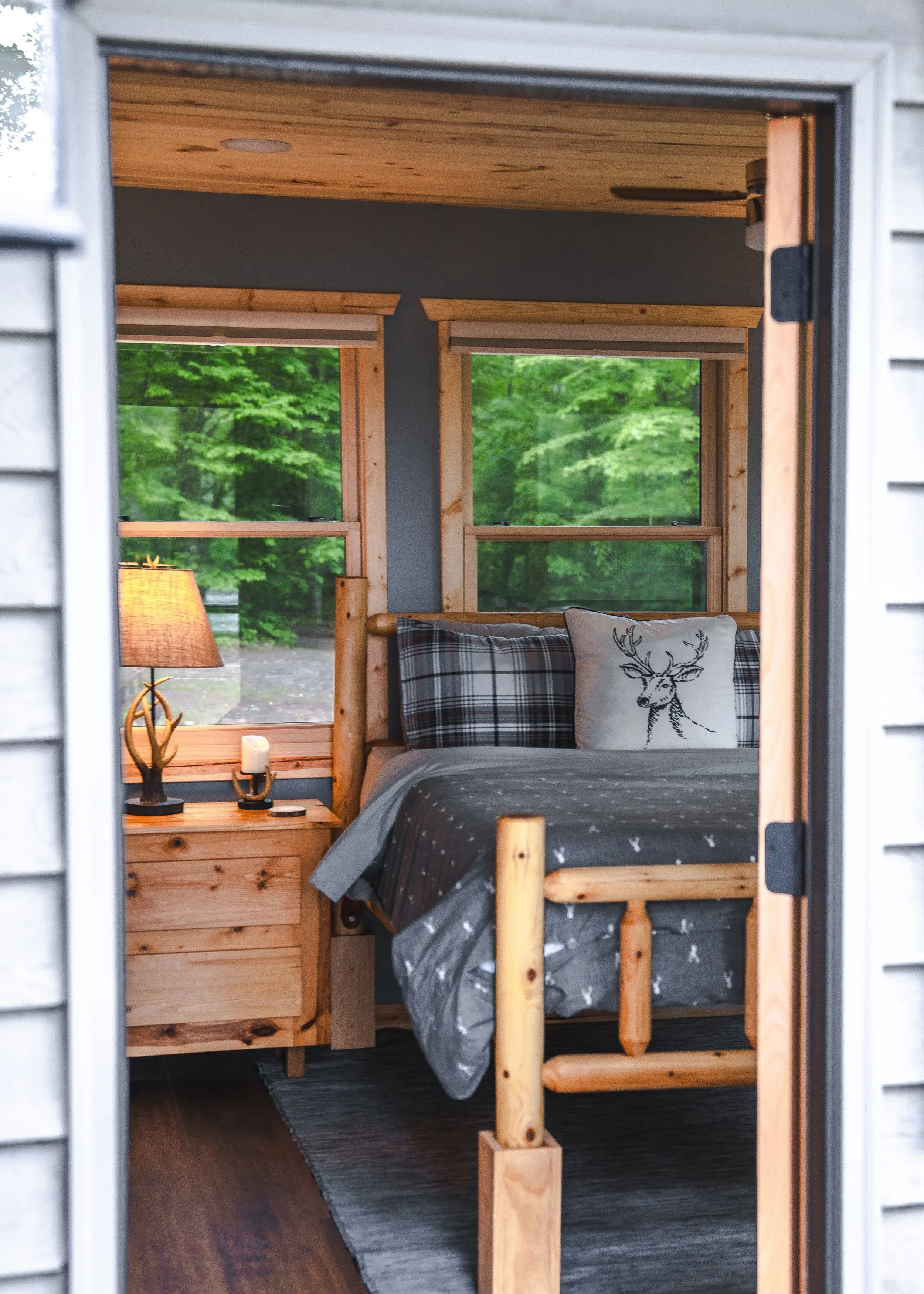 A cozy bedroom with wooden furniture, two windows showing green trees outside, a bed with a deer pillow, plaid and solid pillows, a nightstand with a lamp, a candle, and a small decorative item.
