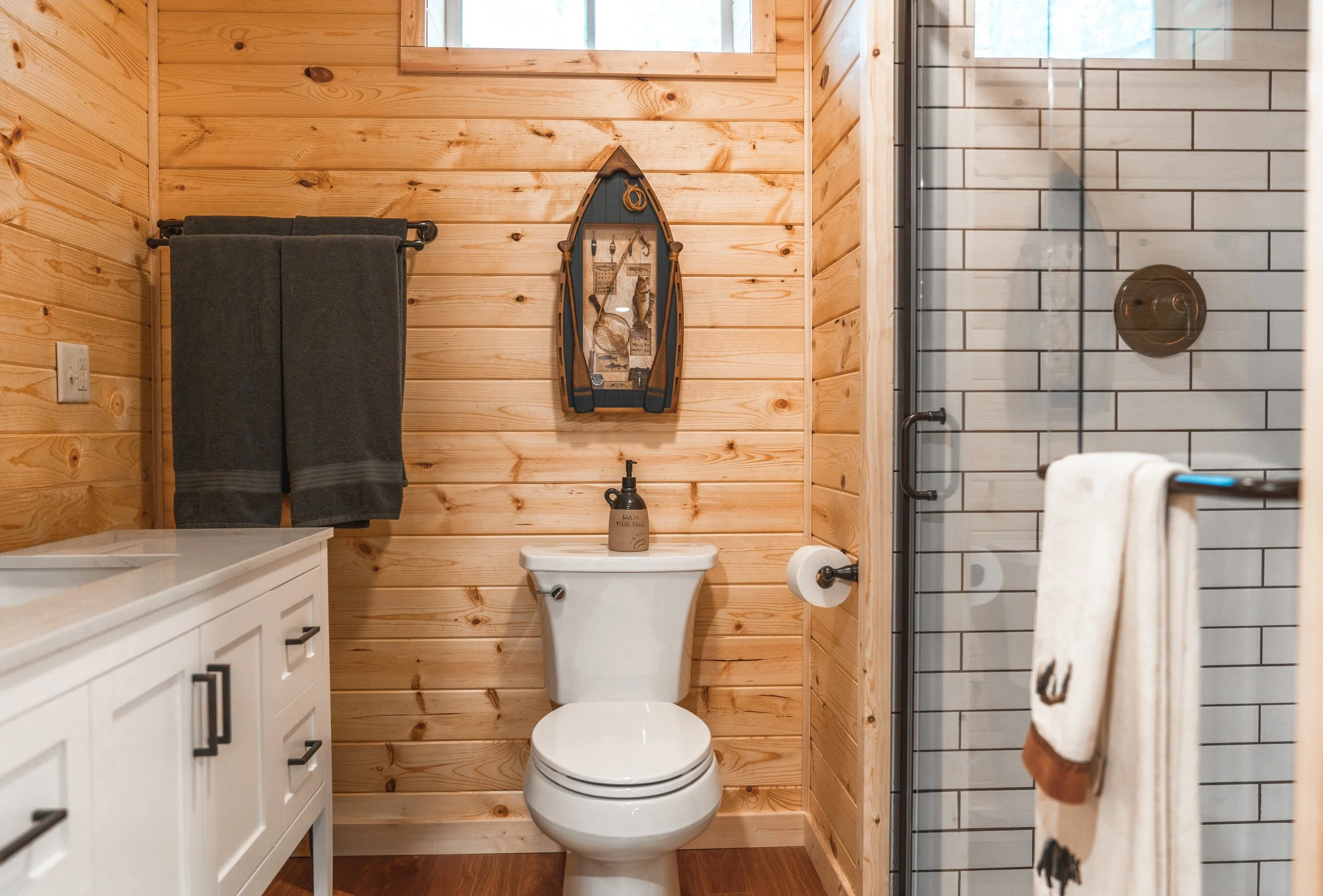 A rustic bathroom with wood-paneled walls, a white toilet, a white cabinet, a wall-mounted towel rack with dark towels, a soap dispenser on the toilet, a decorative boat-shaped shelf with decorations, a glass shower with tiled interior, and a beige towel hanging on a rack.