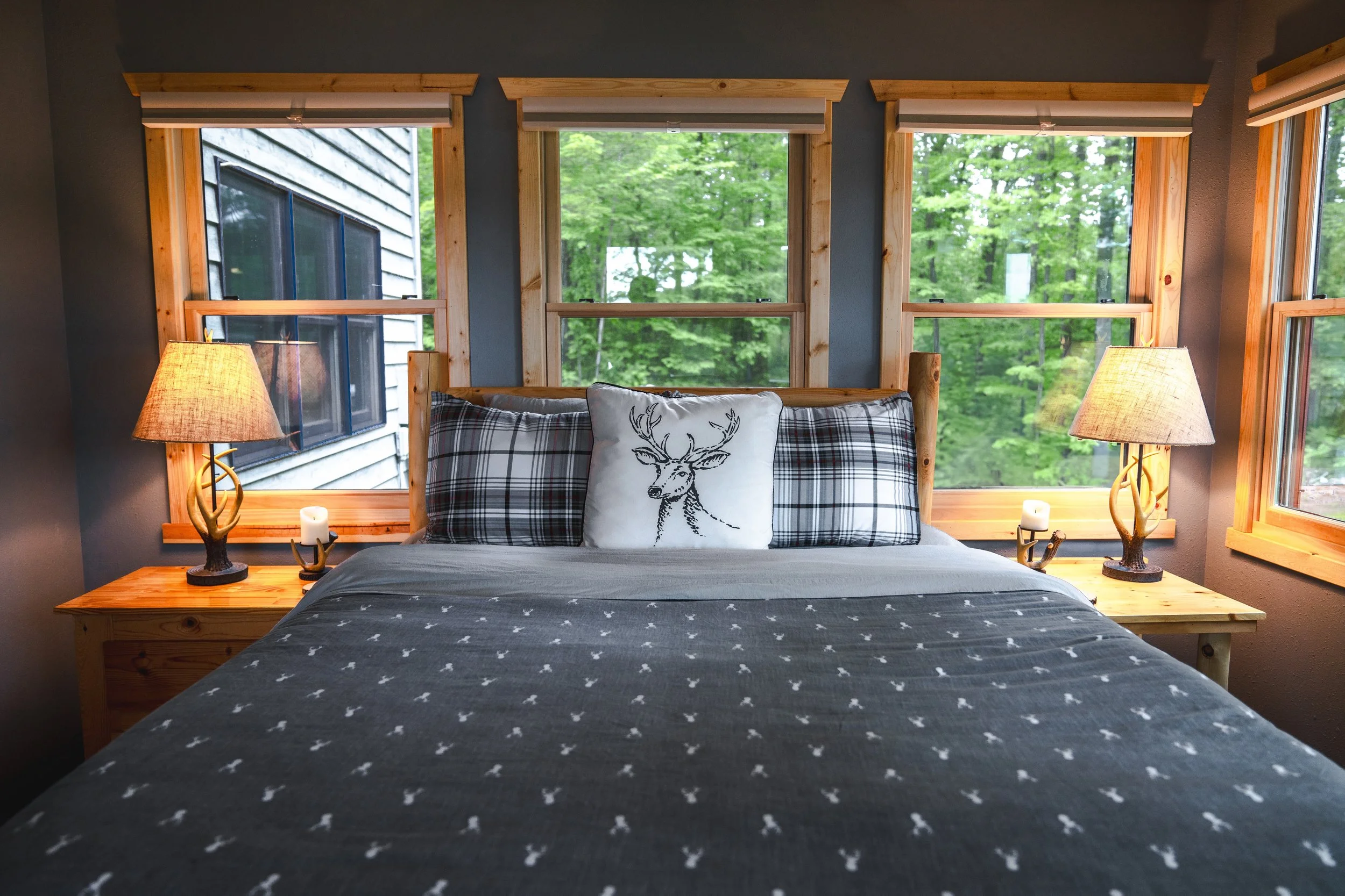 Cozy bedroom with wooden-framed windows, a bed with a deer-illustrated pillow, plaid and solid pillows, and two matching lamps on nightstands, overlooking green trees outside.