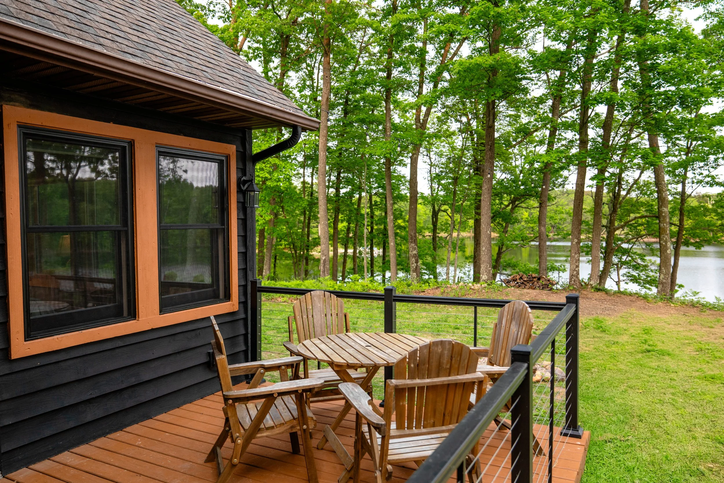 Wooden patio with round table and four chairs, black and brown house exterior with large window, wooded area, lake, and green grass in the background.