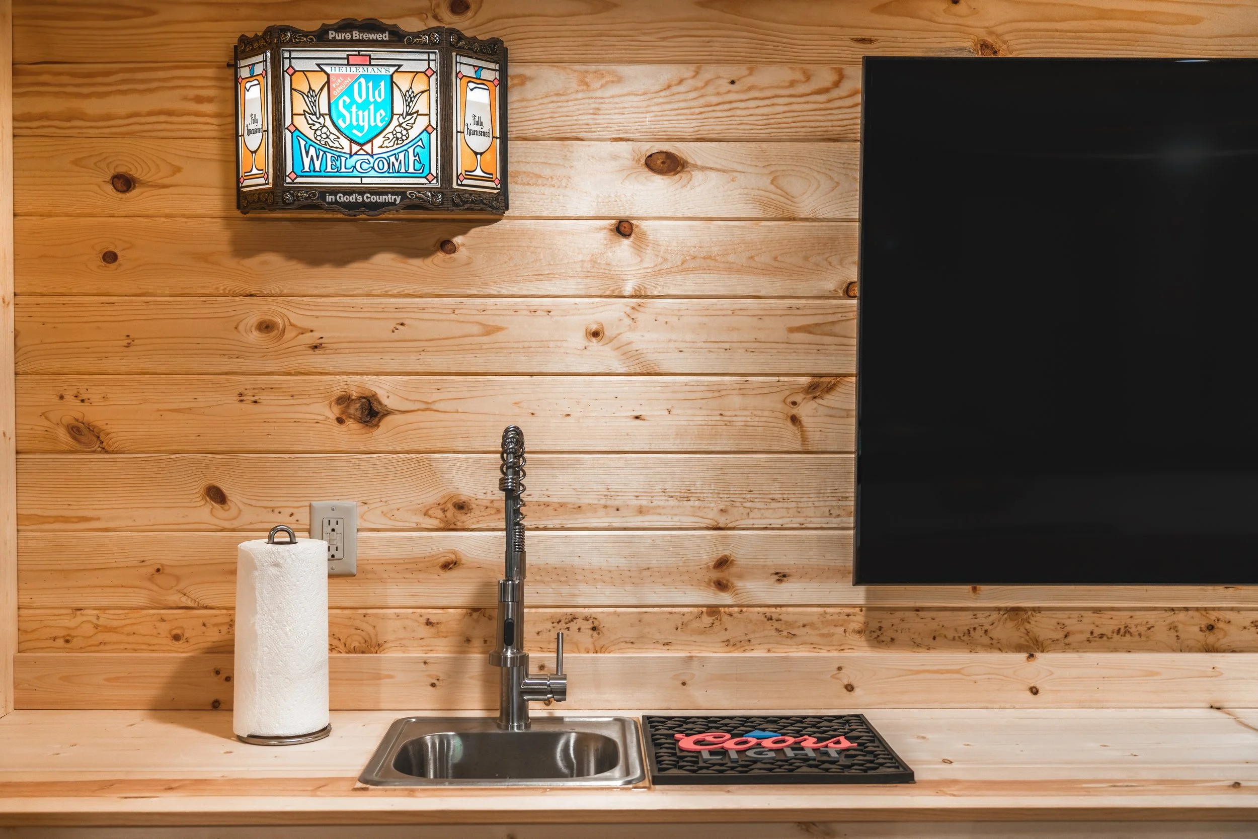 A wooden kitchen counter with a paper towel roll, a stainless steel sink with a tall faucet, a black coasters with red and blue lettering, a flat-screen TV, a wood-paneled wall, and a decorative light with a beer-themed welcome sign.