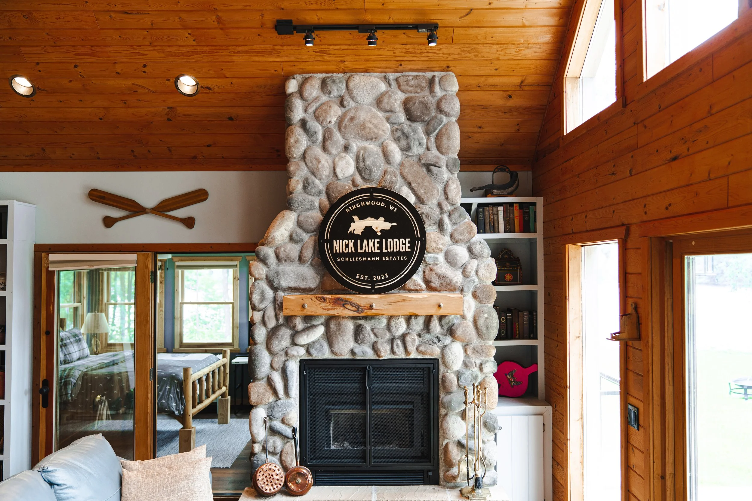 Living room with a large stone fireplace, wooden ceiling, and walls. A sign reads "Nick Lake Lodge, Birchwood, WI, Est. 2023" on the fireplace. There is a shelf with books and decorative items, and a bedroom visible through an open sliding door.