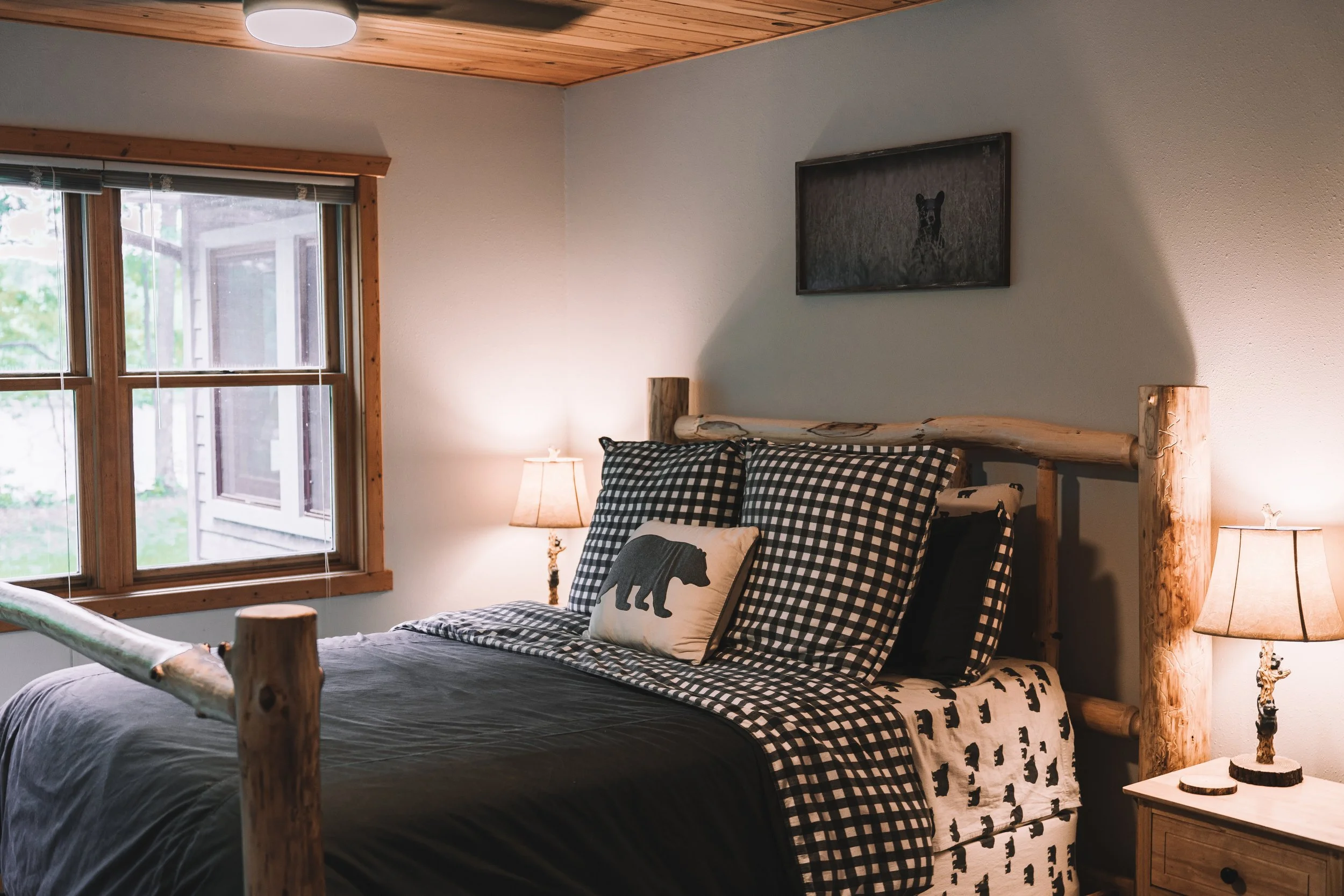 Rustic bedroom with a bed featuring checkered and animal-print bedding, wooden log bed frame, two nightstands with bear-shaped lamps, and a painting of a bear above the bed, near a window with a view of the outdoors.