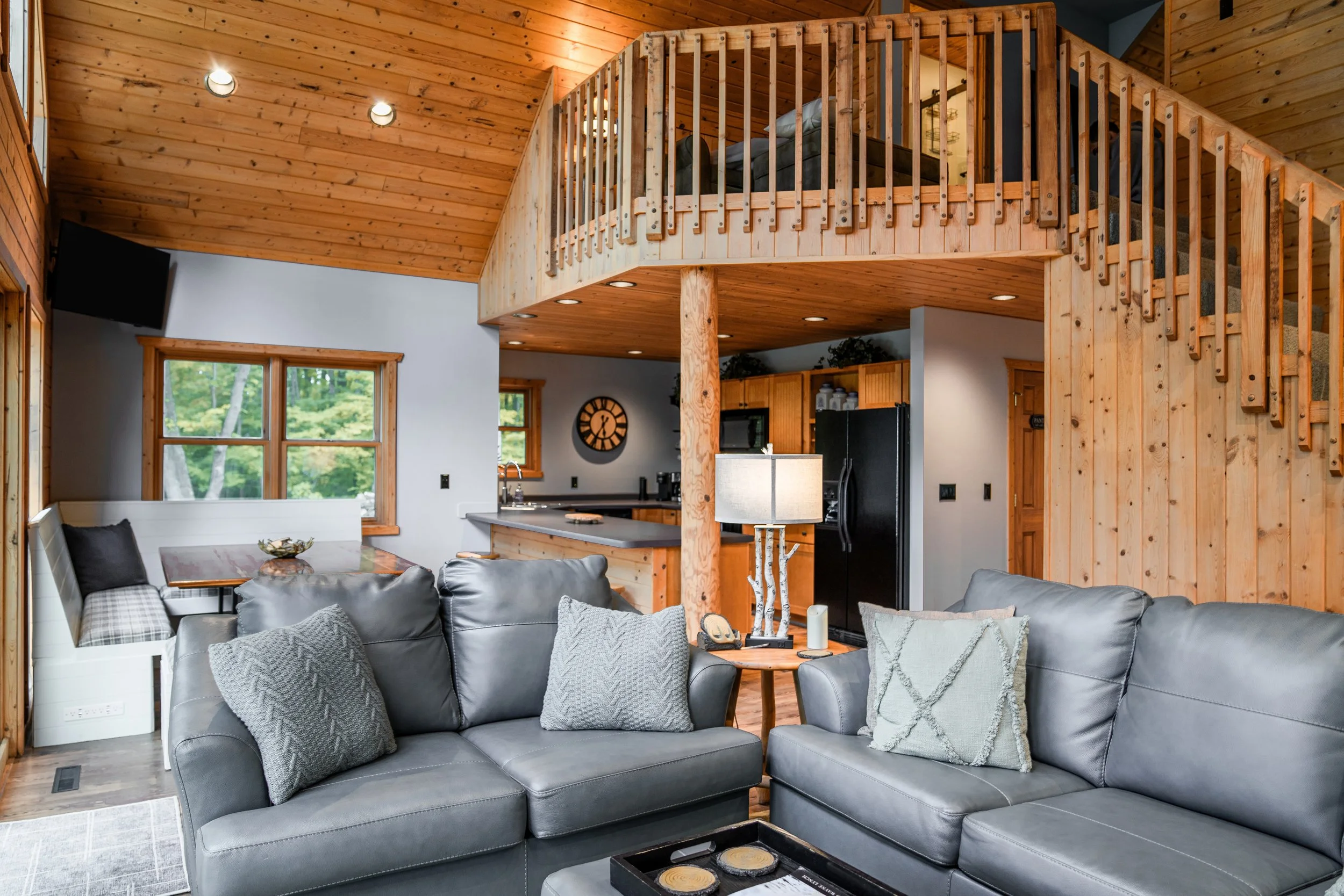 Open concept living room with gray sofas, decorative pillows, a wooden coffee table, and a lofted bedroom with wooden railing, visible from the main living area. The space features wood paneling on the ceiling and walls, large windows with a view of greenery, and a kitchen area in the background.