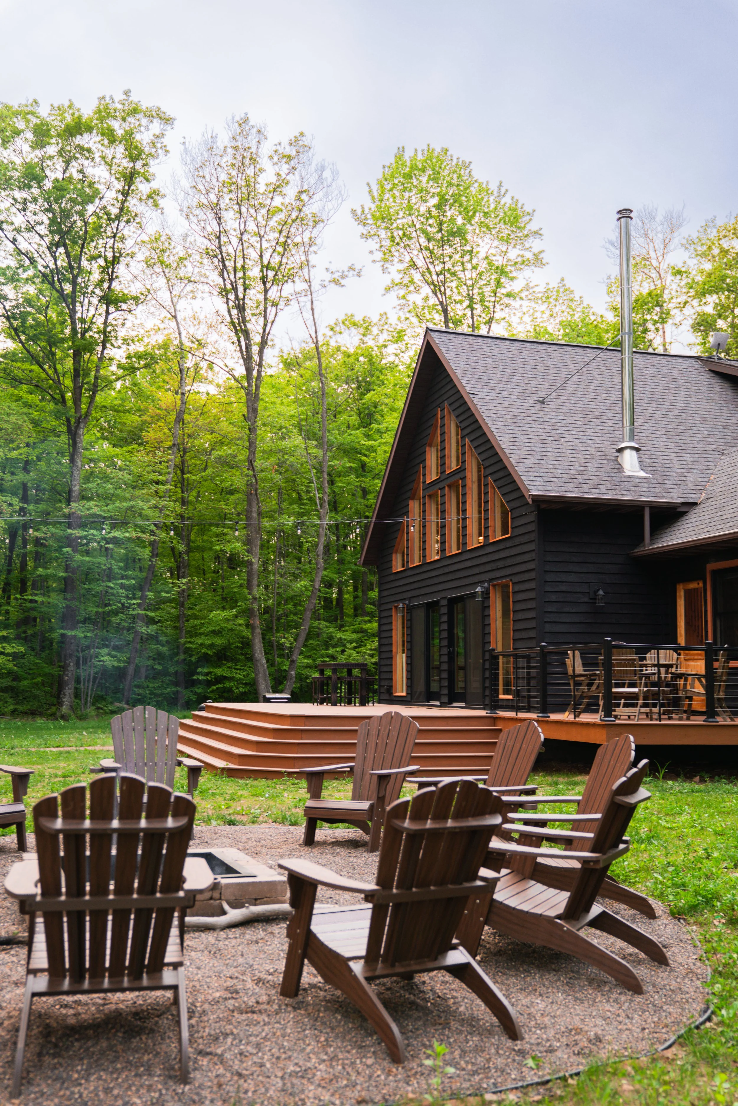 Backyard of a black house with a deck and a fire pit with wooden Adirondack chairs surrounded by trees.