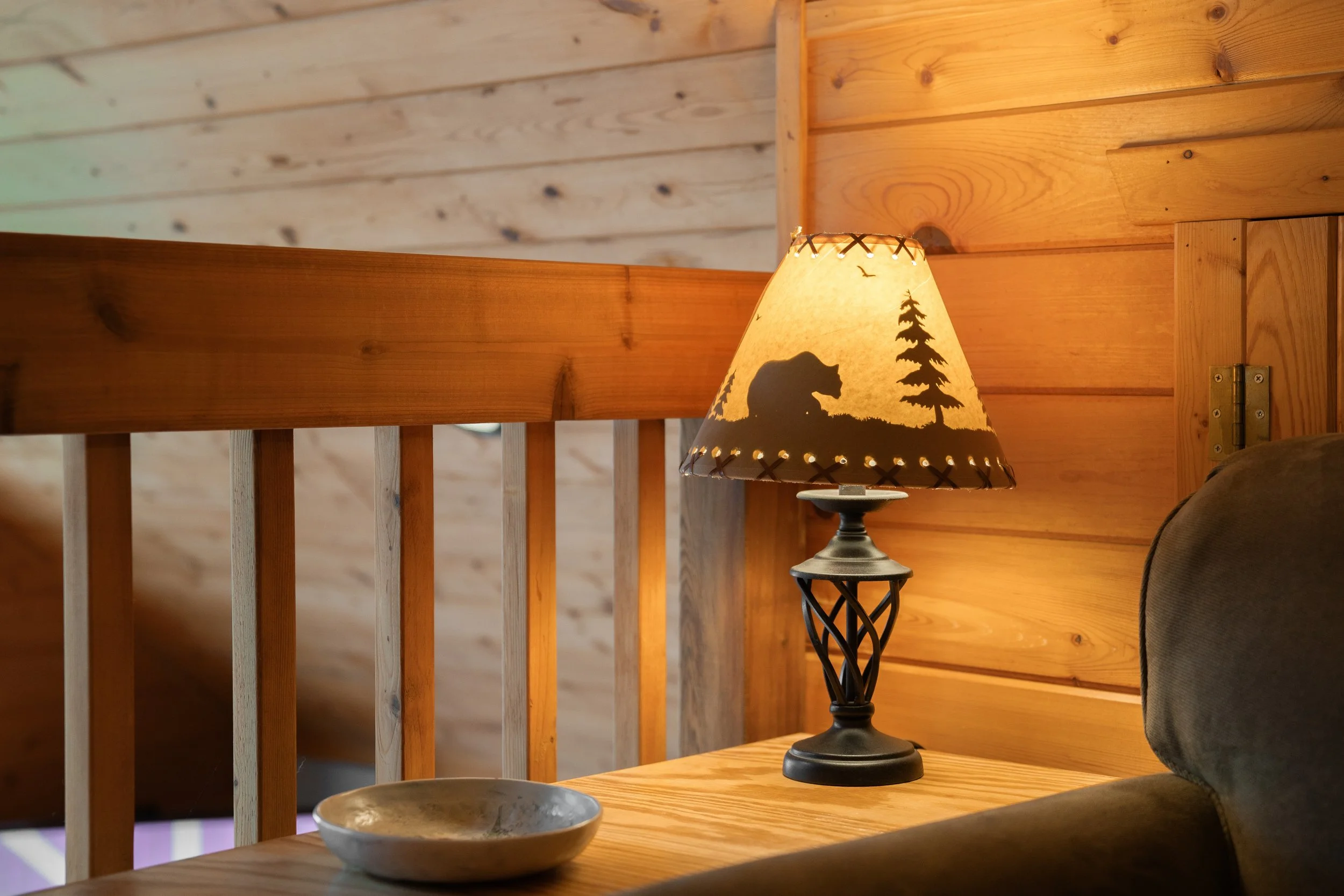 A table lamp with a shade featuring a bear, tree, and bird silhouettes in a cozy wooden cabin interior.