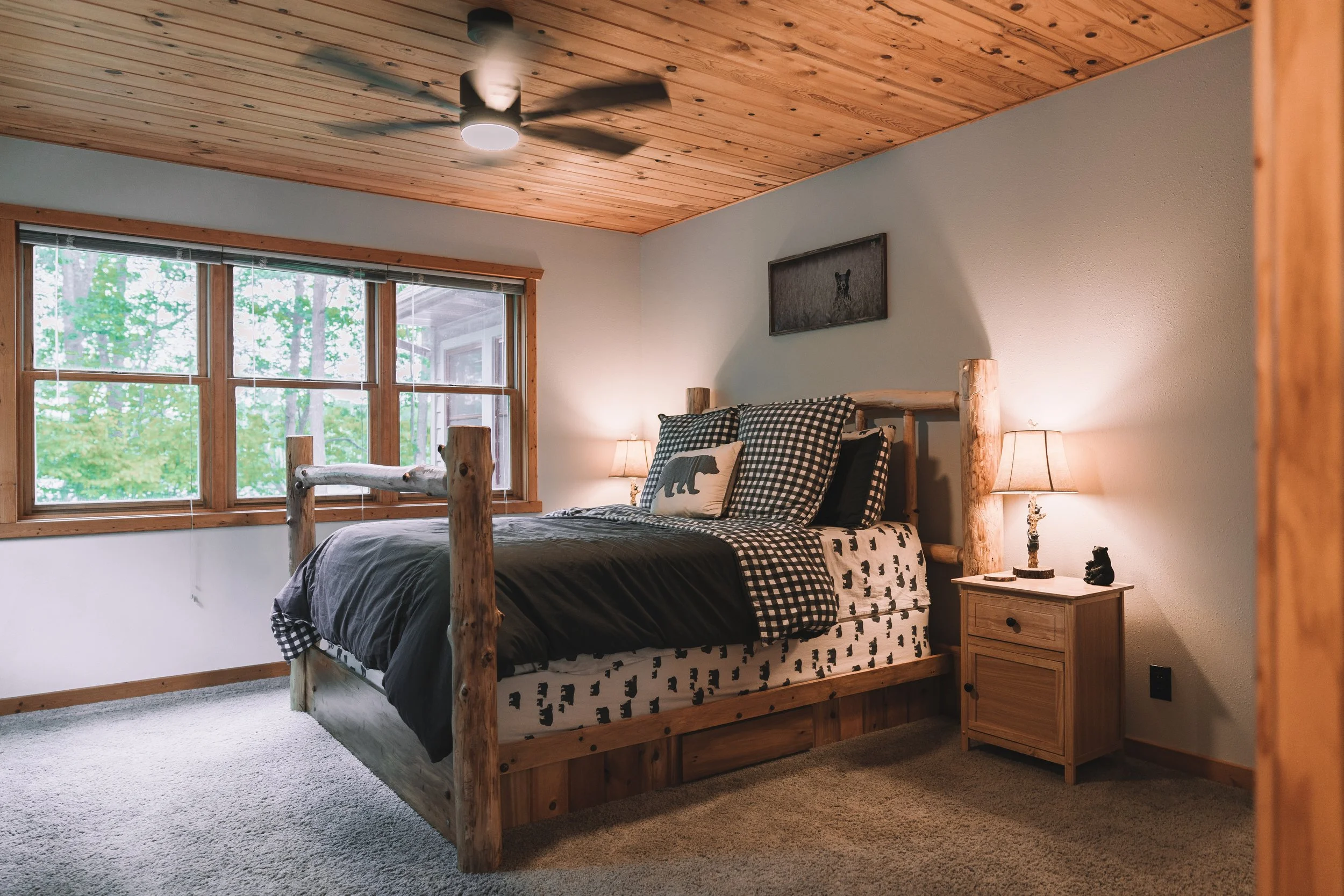 A rustic bedroom with a wooden bed frame, black and white animal-themed bedding, a bedside table with a lamp and decorative bear figurine, and a large window with wooded view.