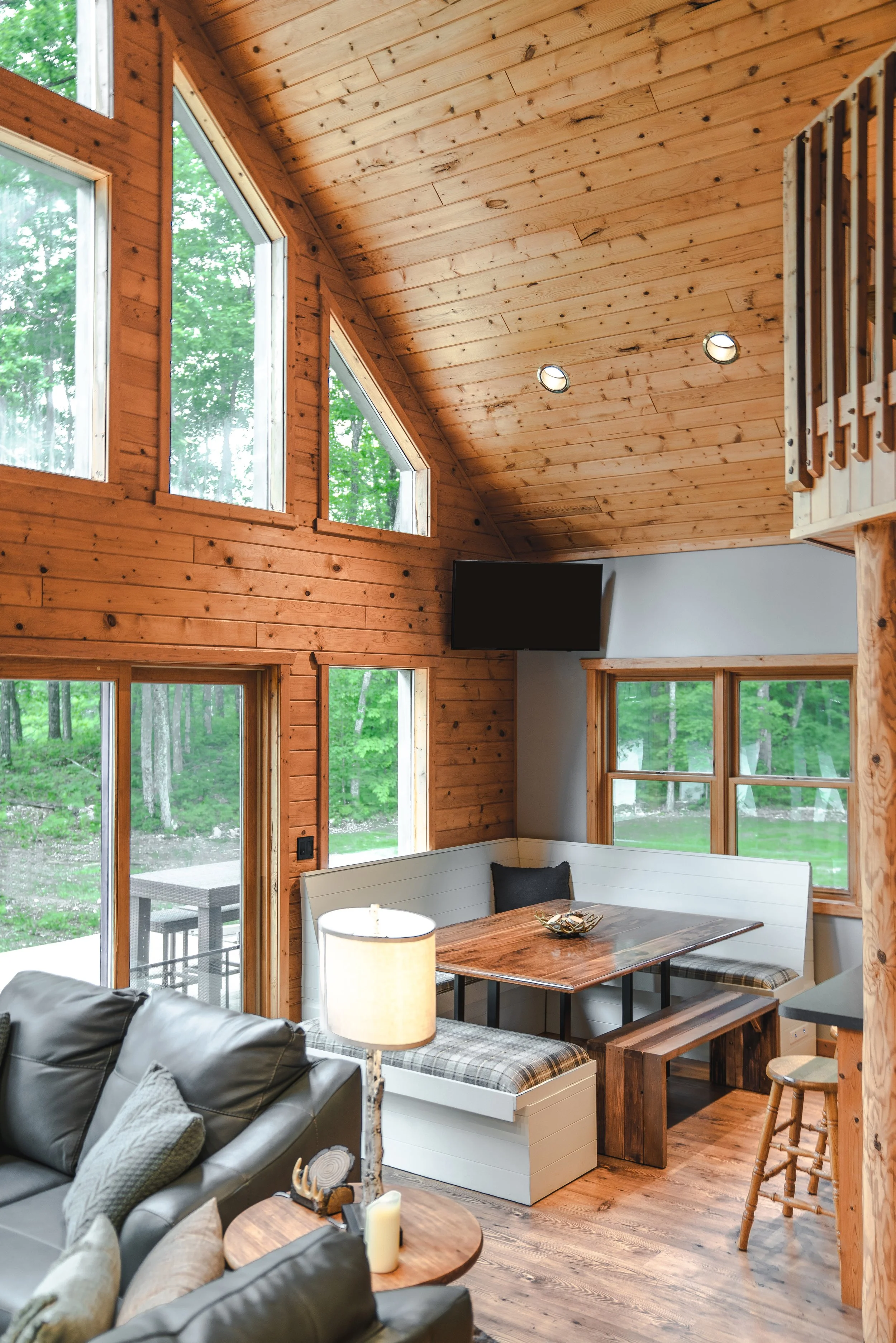Interior of a rustic cabin living room with wood-paneled walls and ceiling, large windows showing greenery outside, a corner nook with a wooden dining table and built-in bench, a black throw pillow on the bench, a television mounted on the wall, and a leather sofa with pillows in the foreground.