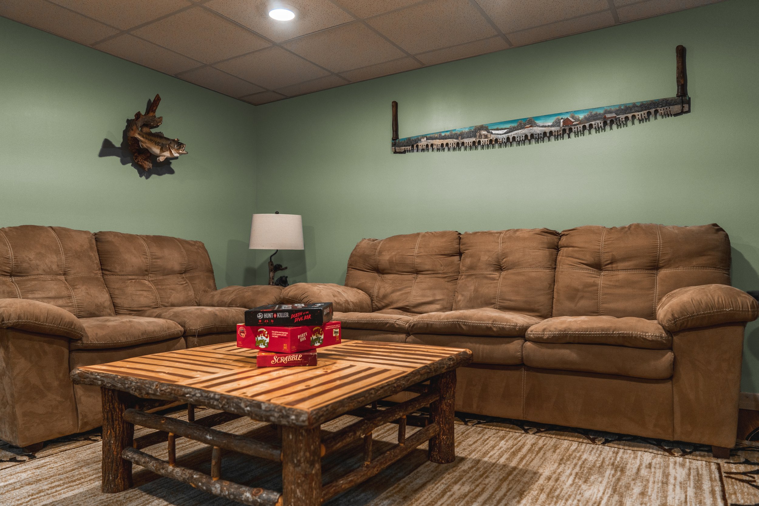 Living room with green walls, two brown sofas, a wooden coffee table with board games, a lamp, a mounted fish on the wall, and a framed artwork.