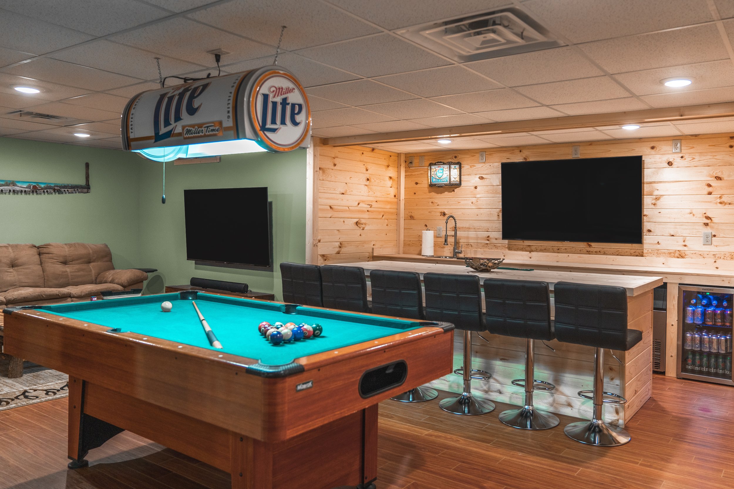 Indoor recreation room with a pool table, a bar counter with four black barstools, a wall-mounted flat-screen TV, a wooden wall paneling, a coin-operated mini-fridge, and a Miller Lite beer sign hanging from the ceiling.