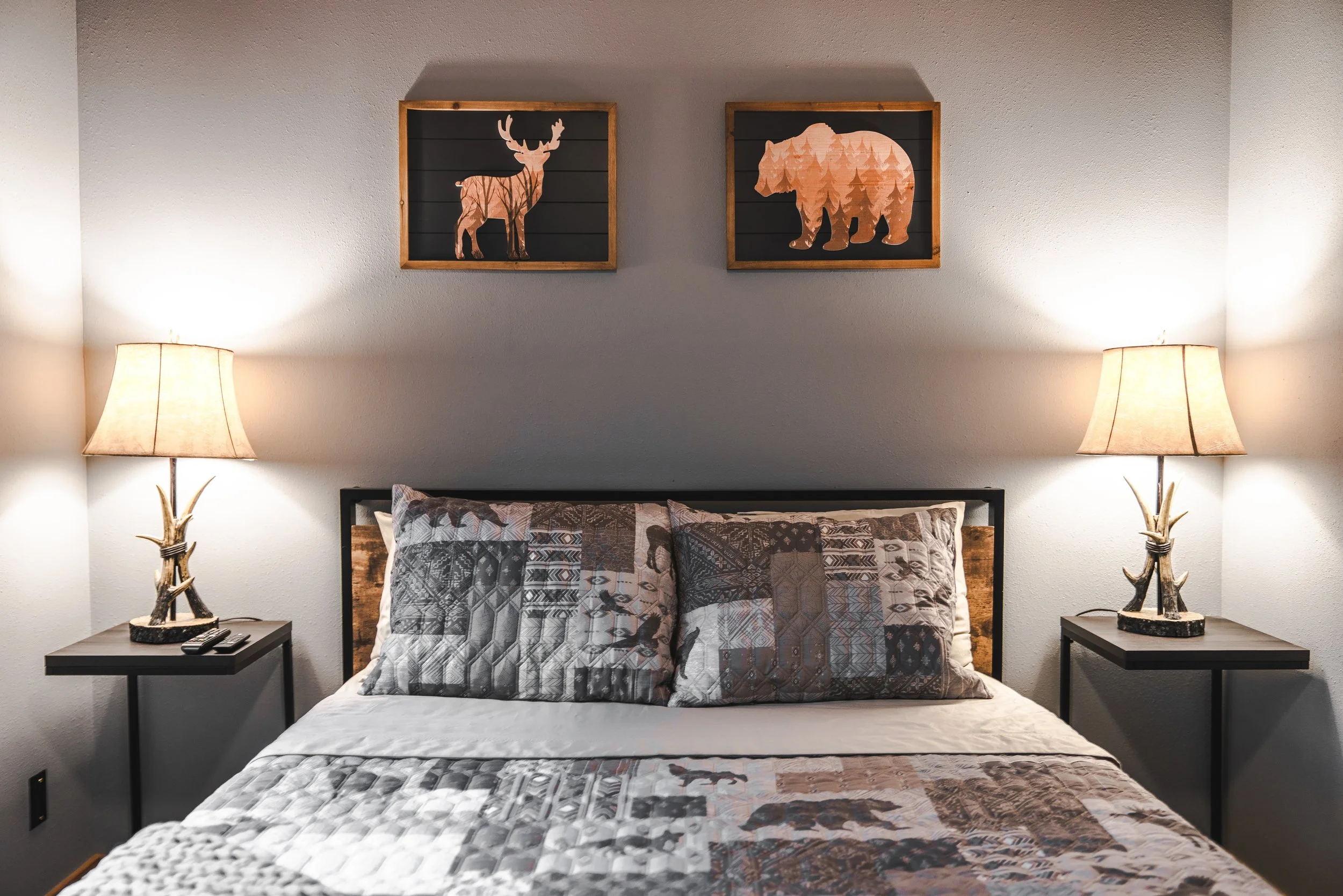 A bedroom with two side tables, each with a lamp with a deer-antler-shaped base, and a bed with a quilted bedspread and pillows. Two framed wall art pieces of a deer and a bear are hung above the bed.