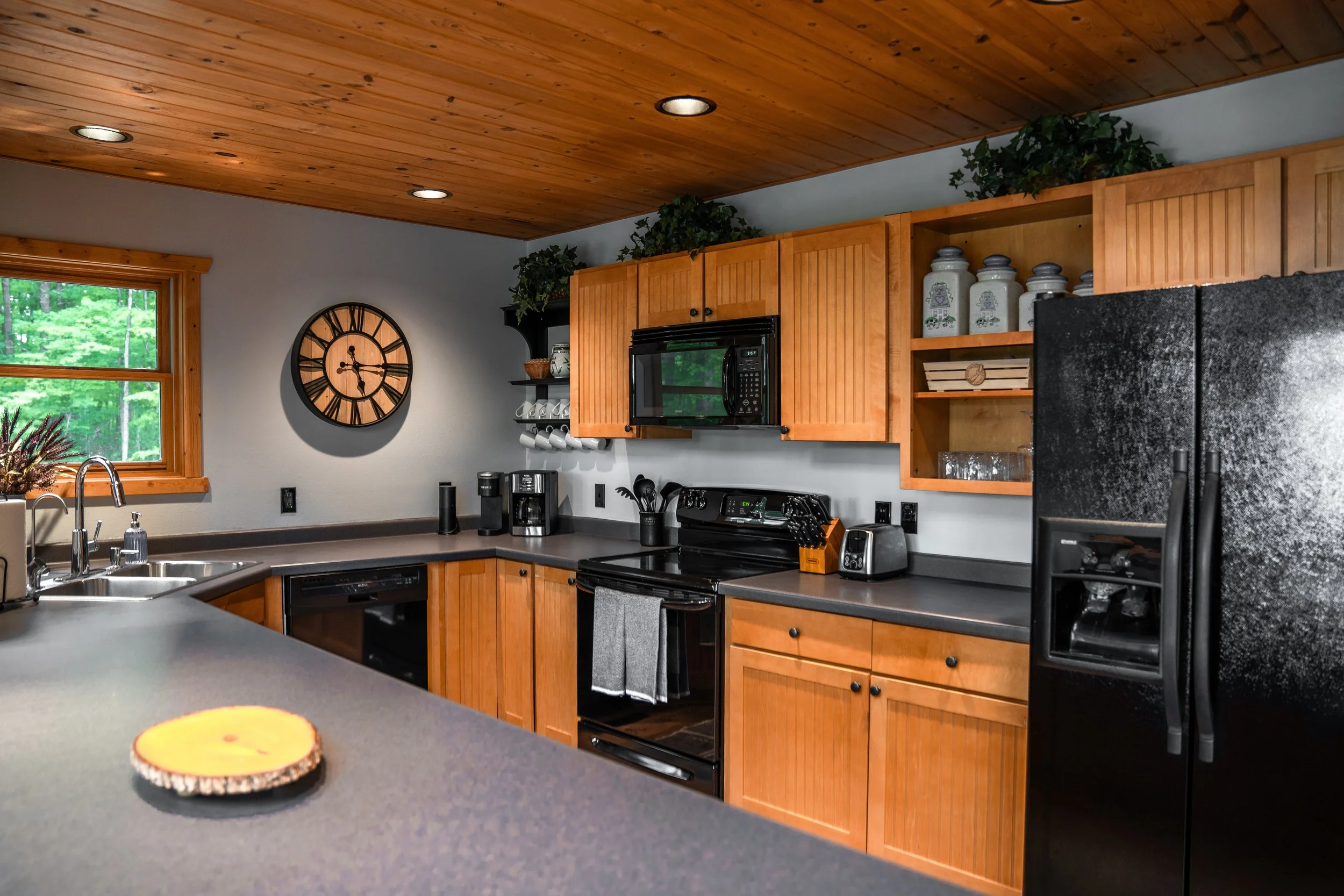 Kitchen with wooden cabinets, black appliances, a gray countertop, a wall clock, a window with green trees outside, and kitchen accessories.