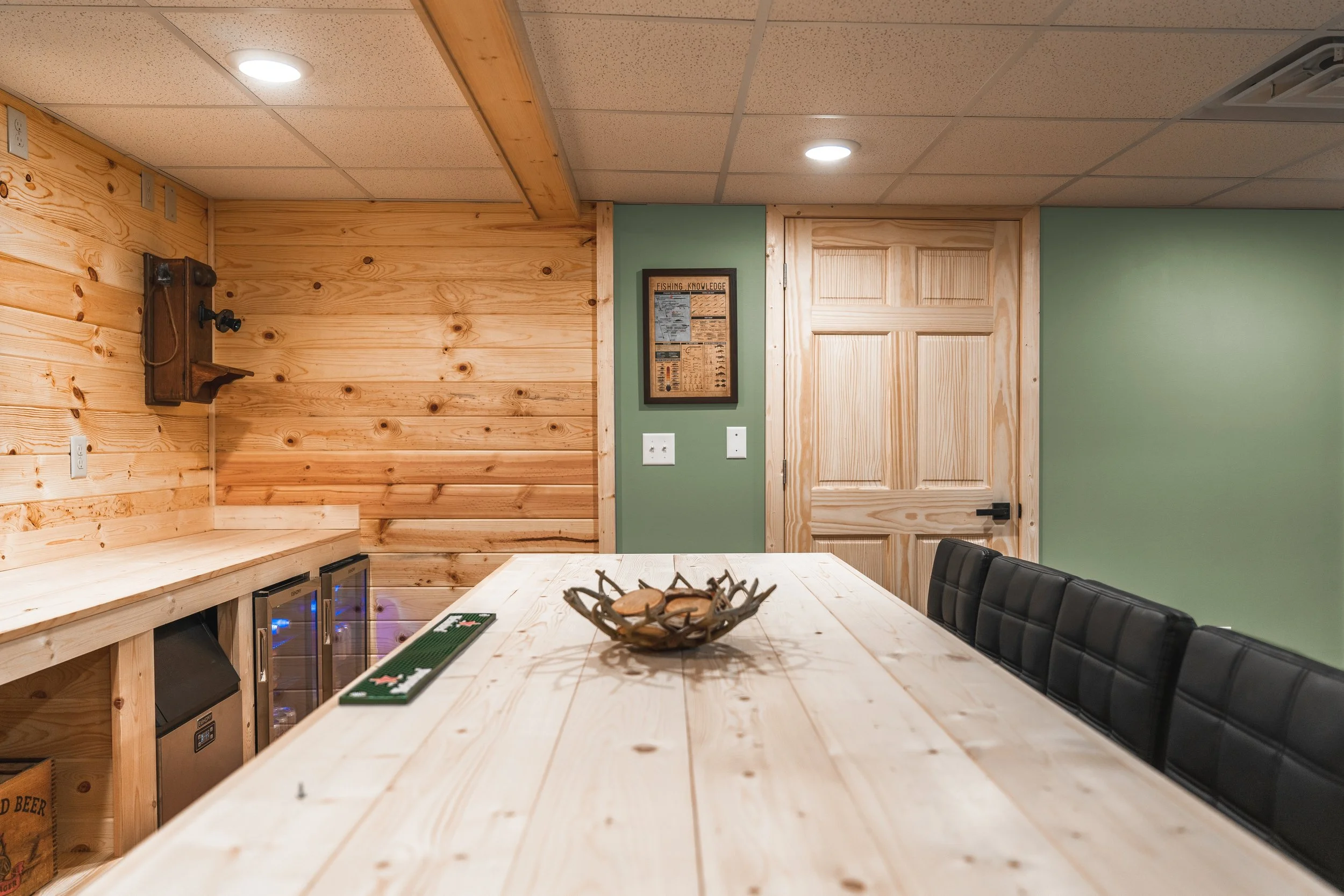 Wood-paneled basement with a large light wood table, black chairs, and a decorative bowl in the center. The room has green and wood walls, a framed poster, and a door. Recessed ceiling lights.