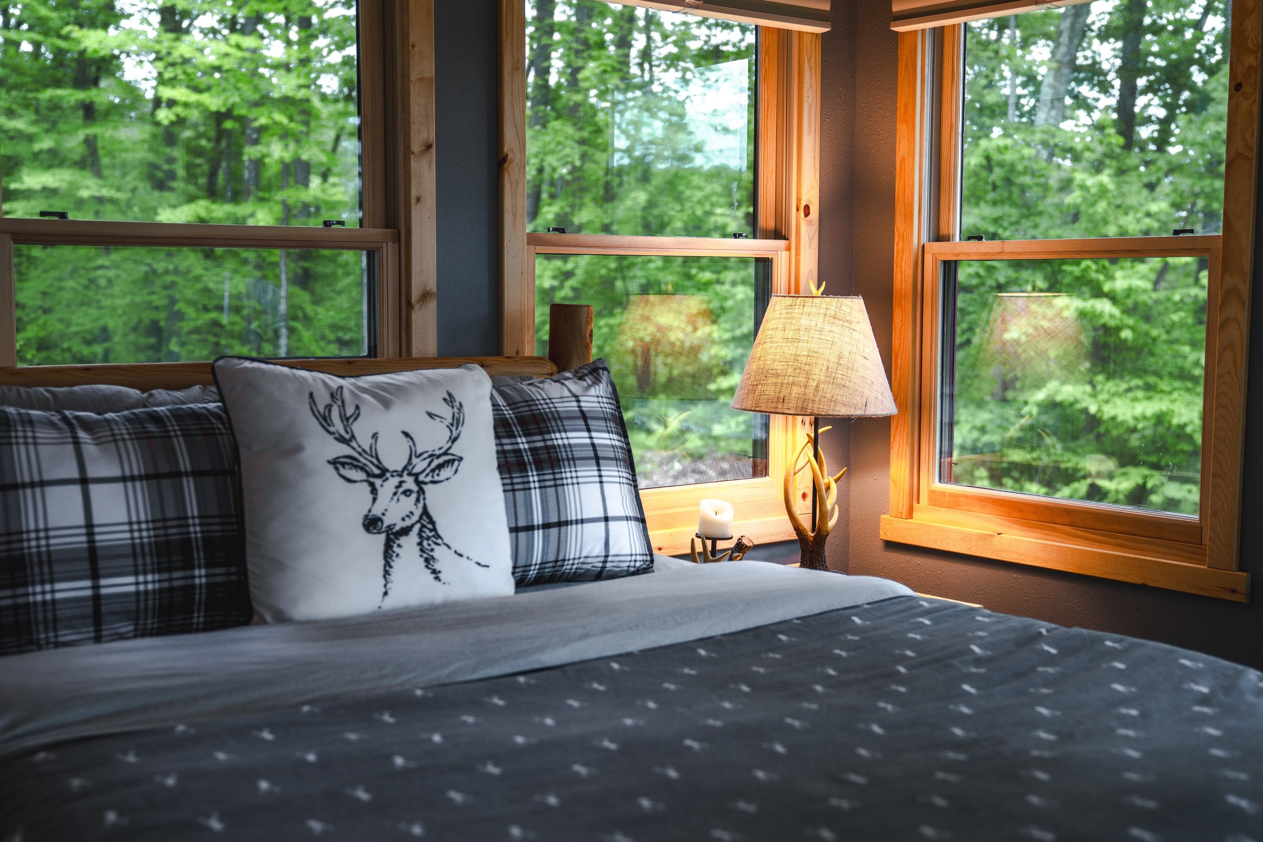 Cozy bedroom with a bed in front of large windows showing a green, wooded landscape outside, decorated with plaid and deer-themed pillows and a lamp with a wooden base.