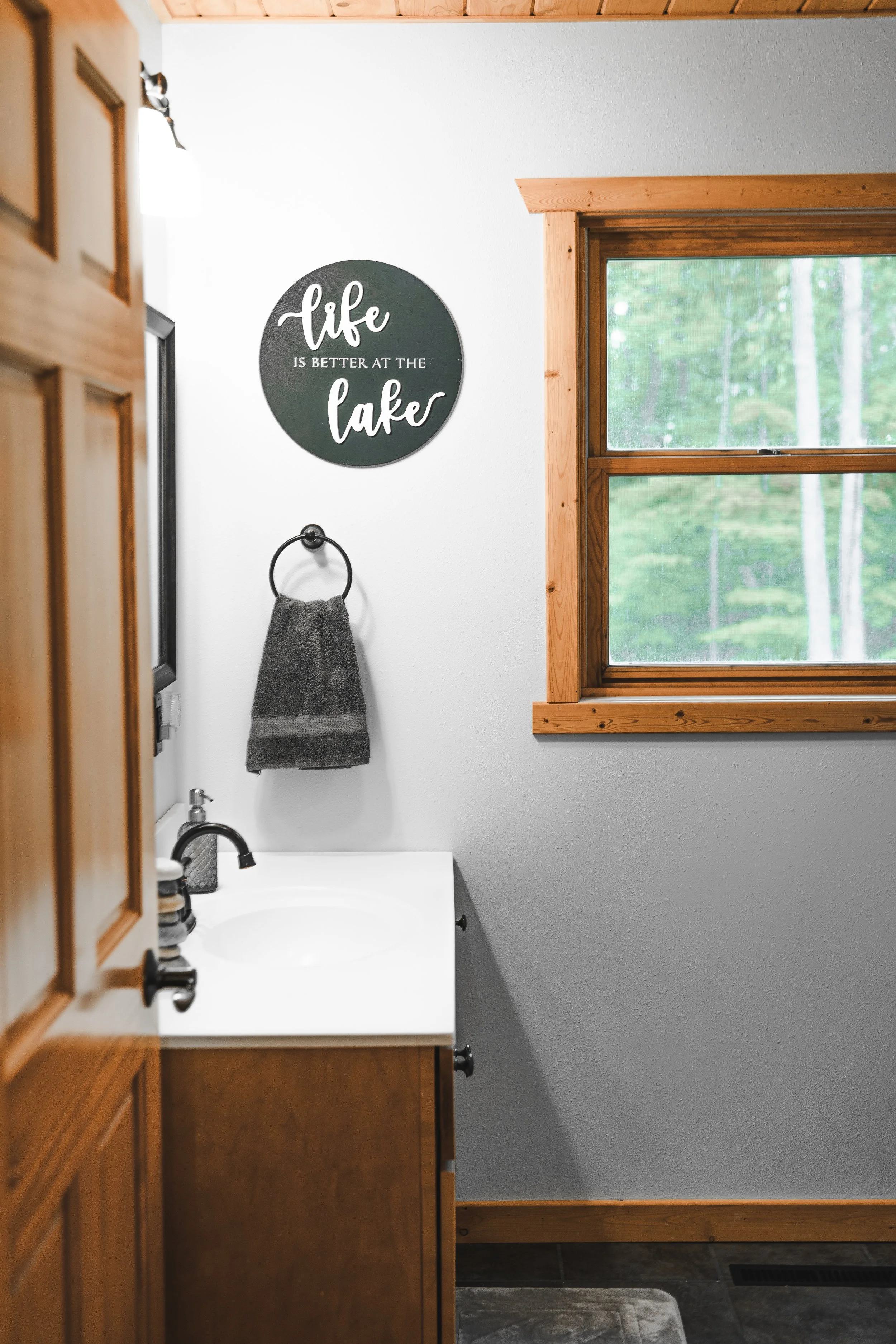 Bathroom with a wooden-framed window, a small white sink with a black faucet, a gray towel on a black ring, and a round sign on the wall that reads 'life is better at the lake'