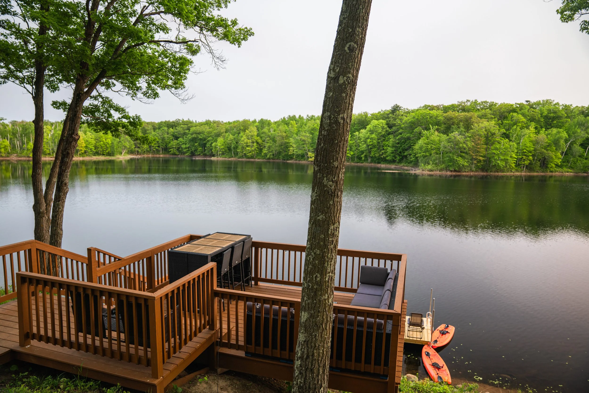 A wooden deck overlooking a calm lake surrounded by lush green trees, with two orange kayaks docked at the water's edge.